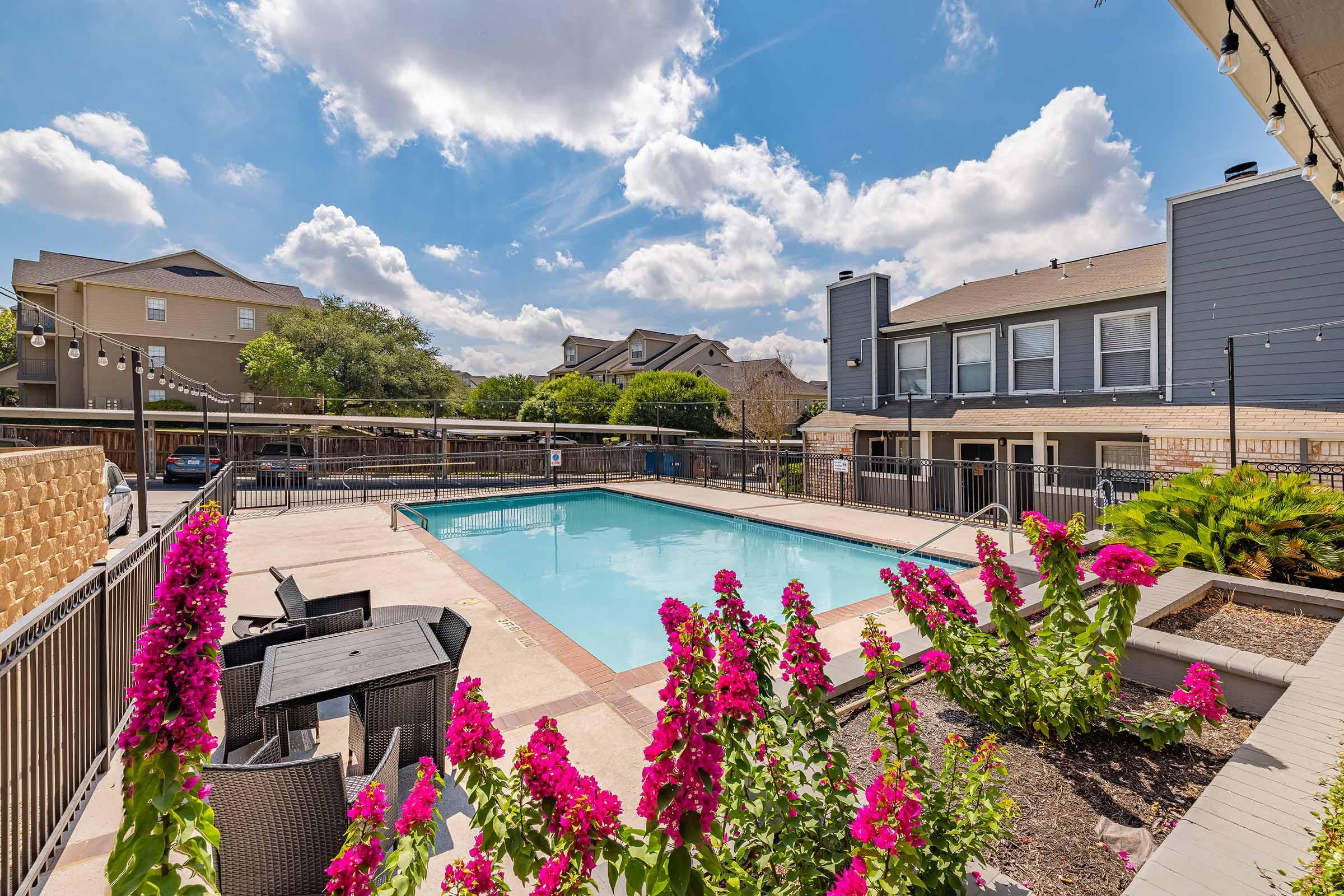 A clear swimming pool surrounded by a fenced area, with vibrant pink flowers in the foreground. The scene features houses in the background under a blue sky with fluffy clouds. Outdoor seating is visible near the pool area, creating a relaxing atmosphere.