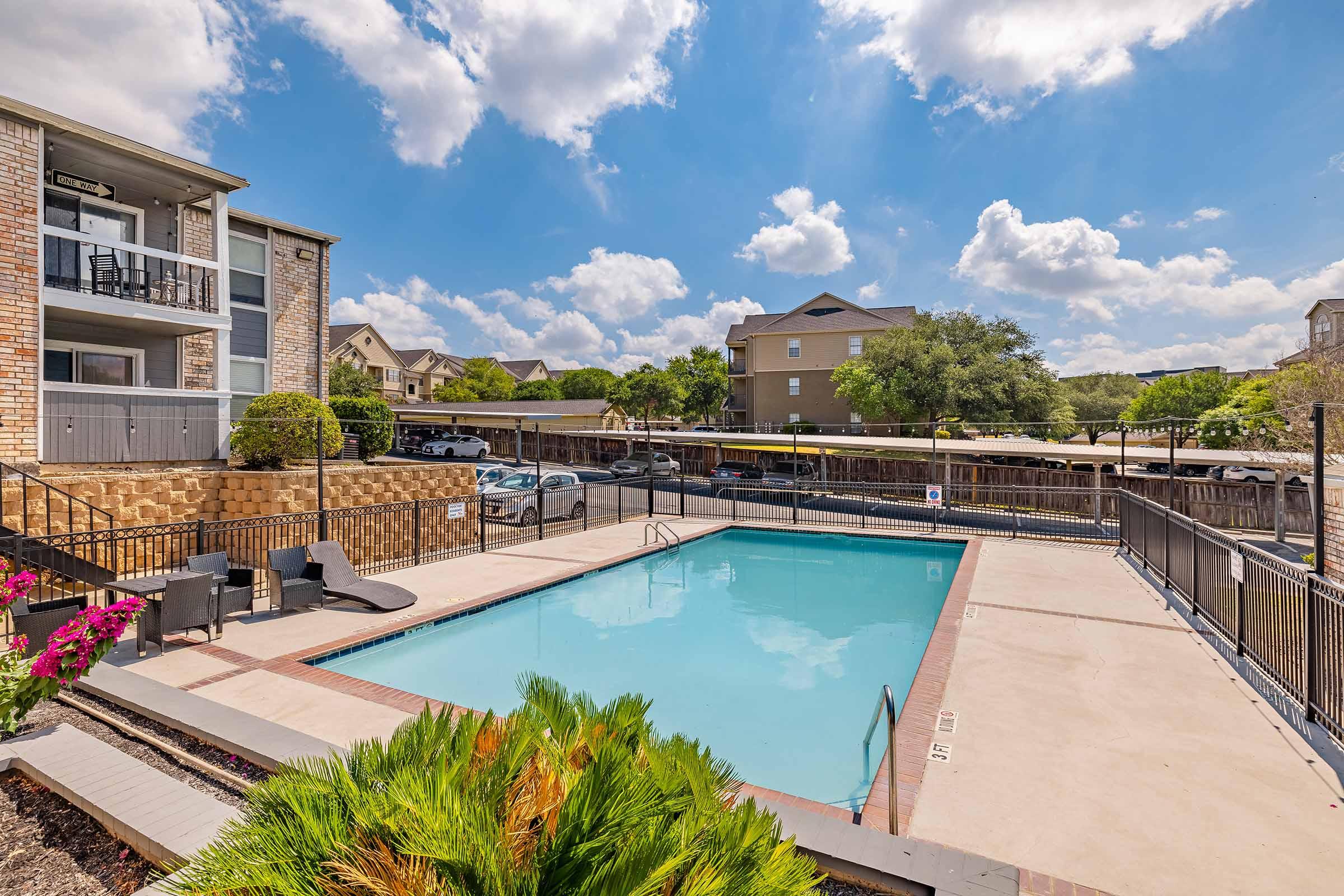 A bright outdoor pool surrounded by a patio area, with lounge chairs and blooming flowers in the foreground. Apartment buildings are visible in the background under a partly cloudy sky. The scene suggests a relaxing and inviting atmosphere for residents and guests.
