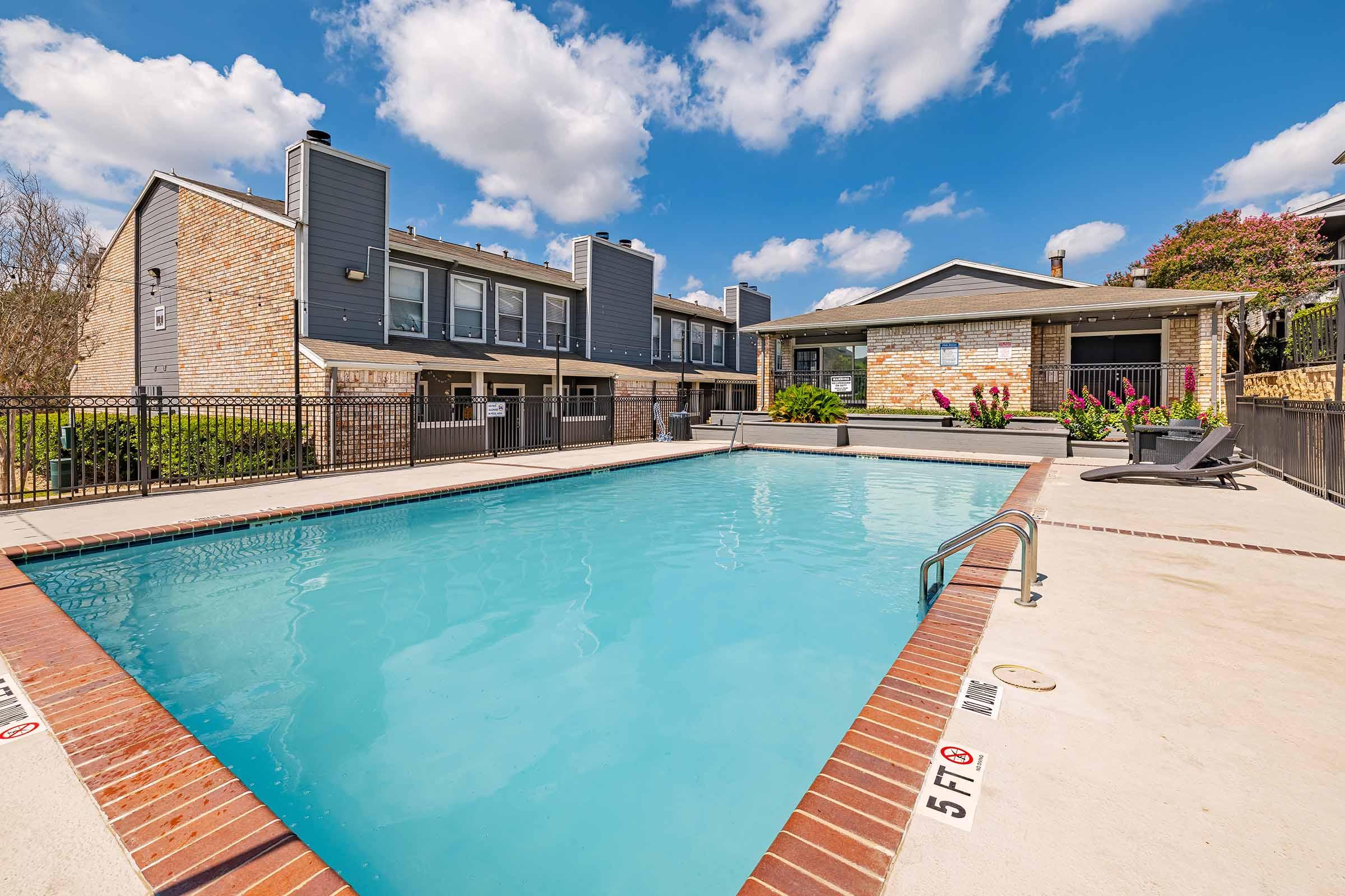 A clean, inviting swimming pool surrounded by a well-maintained patio area. In the background, modern apartment buildings with gray and brick exteriors, complemented by lush greenery and colorful flowers. Bright blue skies with fluffy white clouds enhance the serene atmosphere.