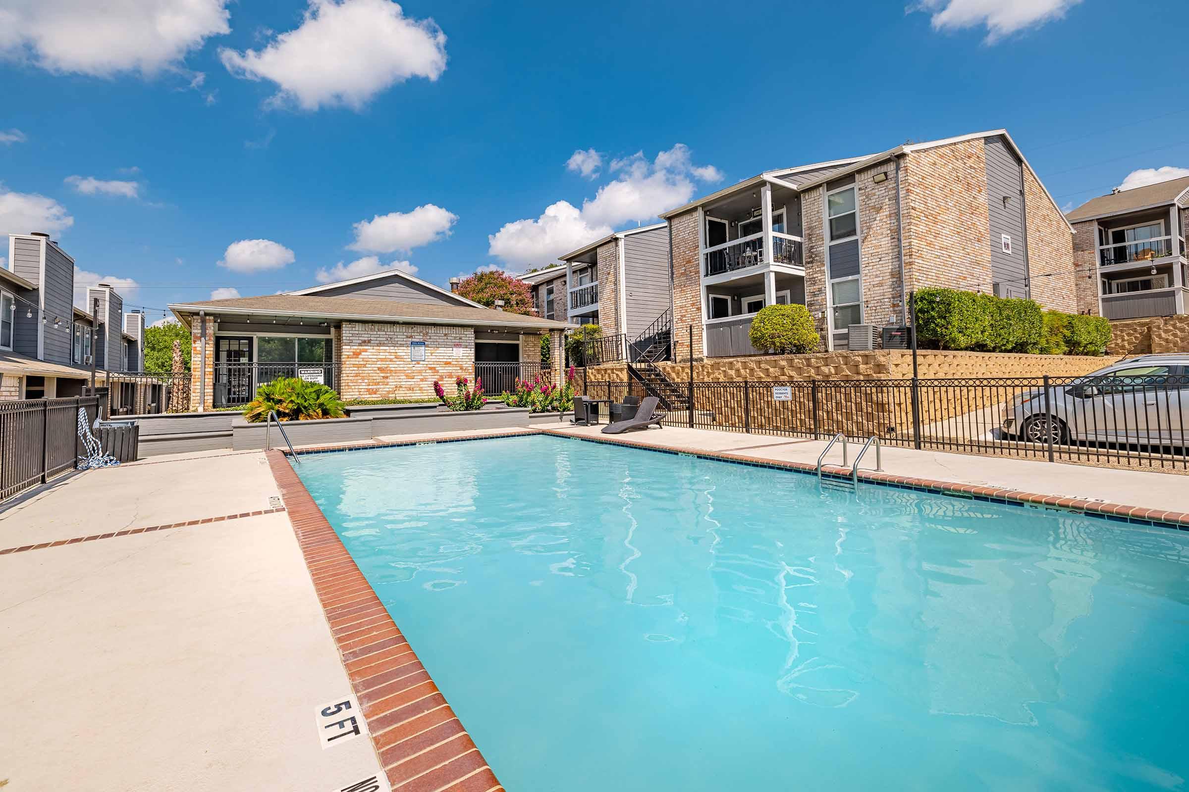 A brightly lit apartment complex pool area featuring a clear blue swimming pool surrounded by a patio. A small building with large windows serves as a poolside clubhouse, while well-maintained landscaping adds color. The setting is under a clear blue sky with a few fluffy clouds.