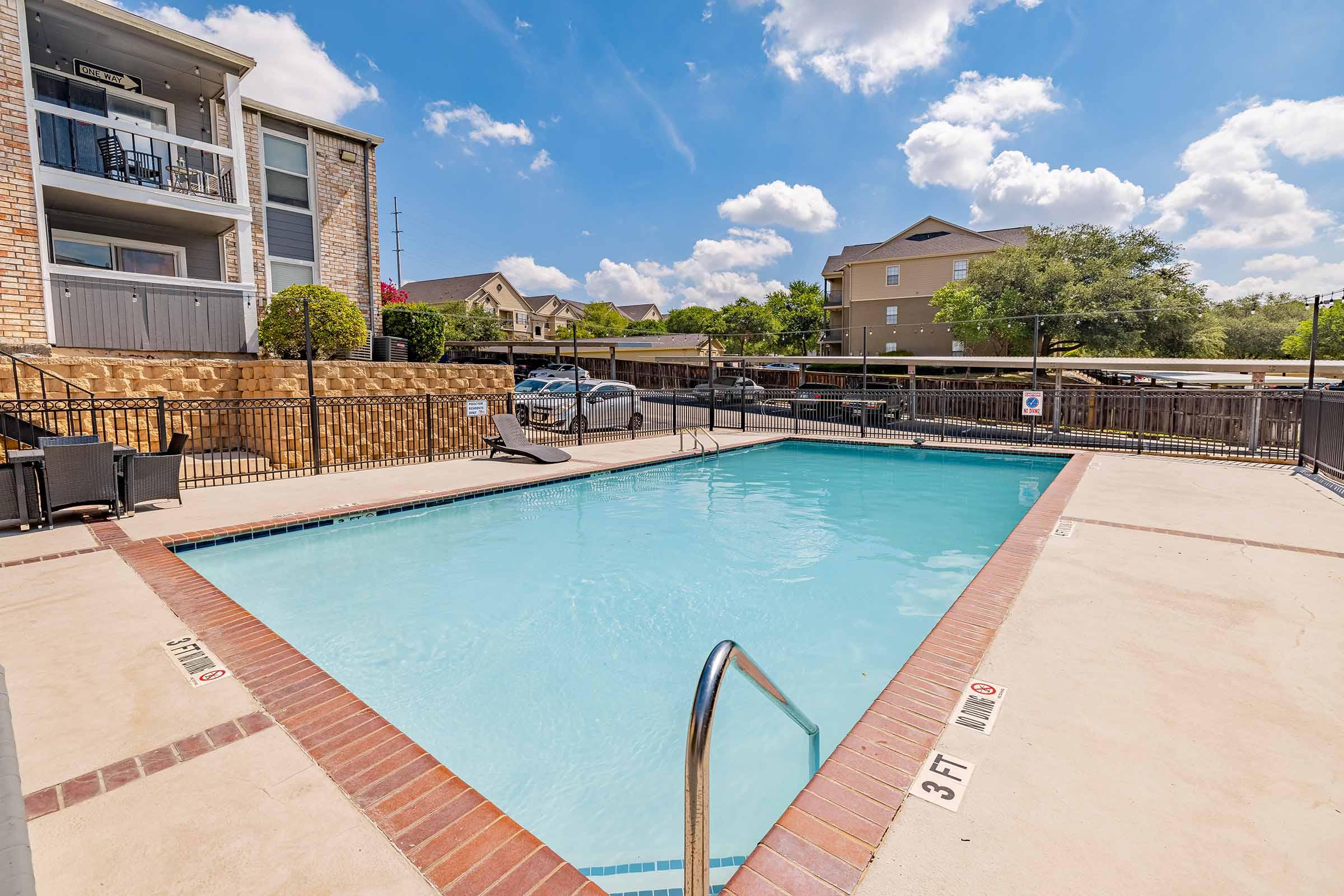 A clean, inviting swimming pool surrounded by a patio area. The pool features a ladder for entry and is marked with a depth sign. In the background, there are residential buildings and a parking area. The sky is bright with fluffy white clouds.