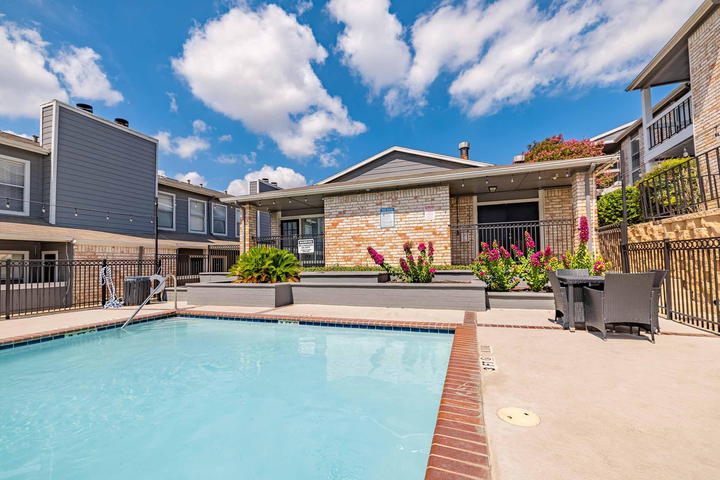 A bright outdoor scene featuring a swimming pool in the foreground, surrounded by a sunlit patio. In the background, there is a brick house with large windows and colorful flowering plants. The sky is partly cloudy, adding a pleasant ambiance to the setting.