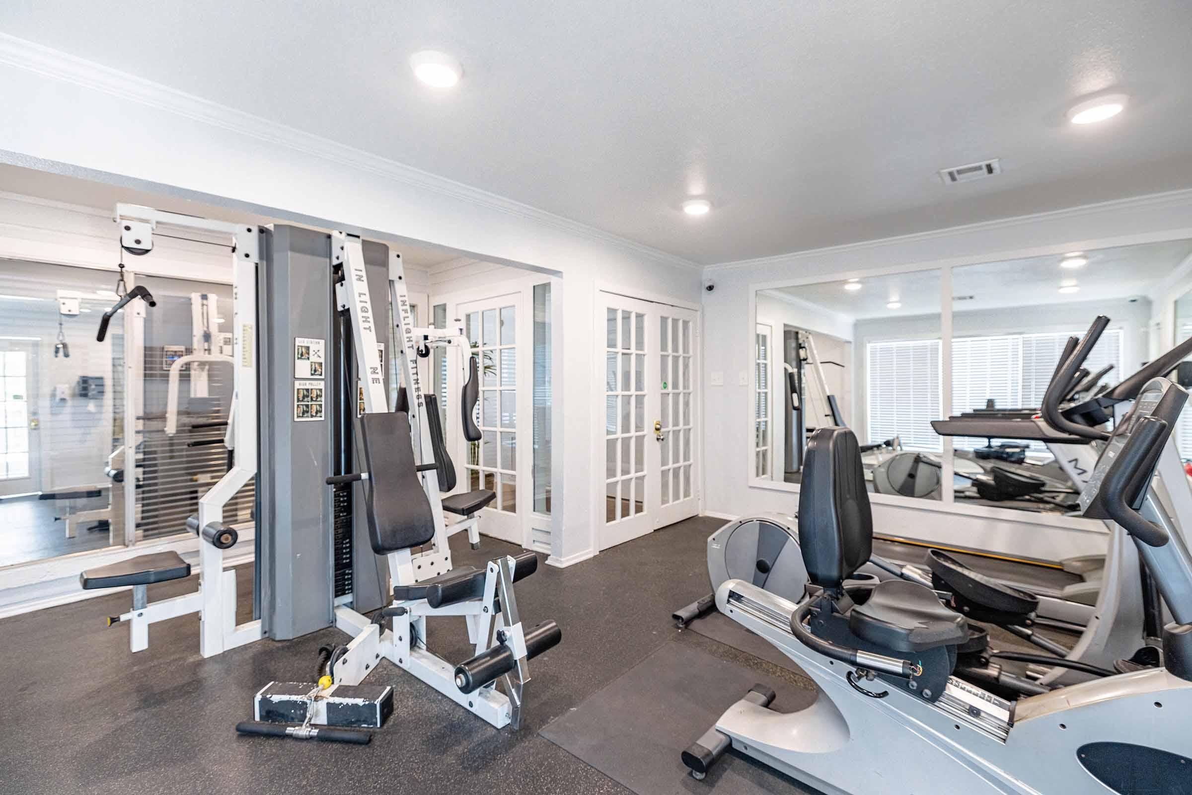 A spacious home gym featuring various exercise equipment, including a multi-station weight machine, stationary bikes, and a treadmill. Large mirrors reflect the equipment, and bright lighting enhances the open feel of the space. The flooring is dark, contrasting with the light-colored walls and natural light from windows.