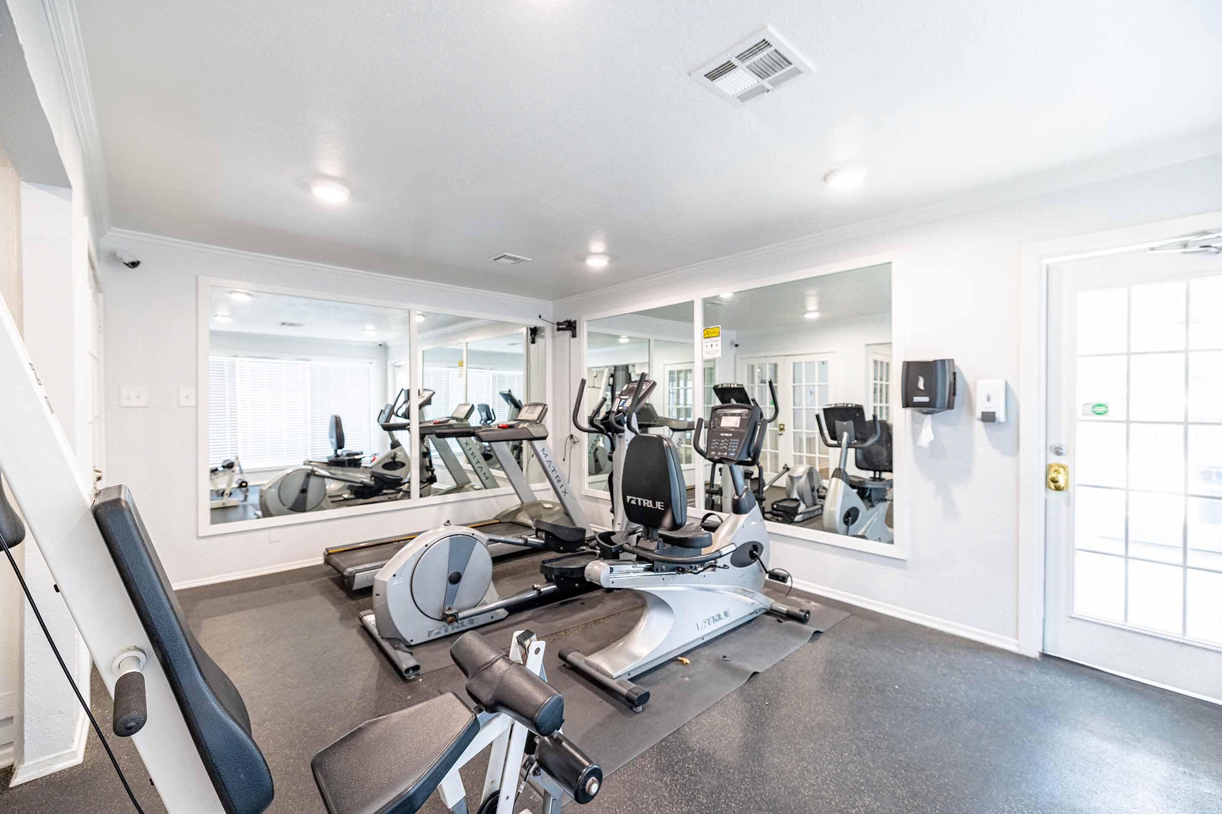 A modern gym interior featuring exercise machines including treadmills and stationary bikes, large mirrors reflecting the workout area, and bright lighting. The room has a clean, organized appearance with a door leading outside visible in the background.