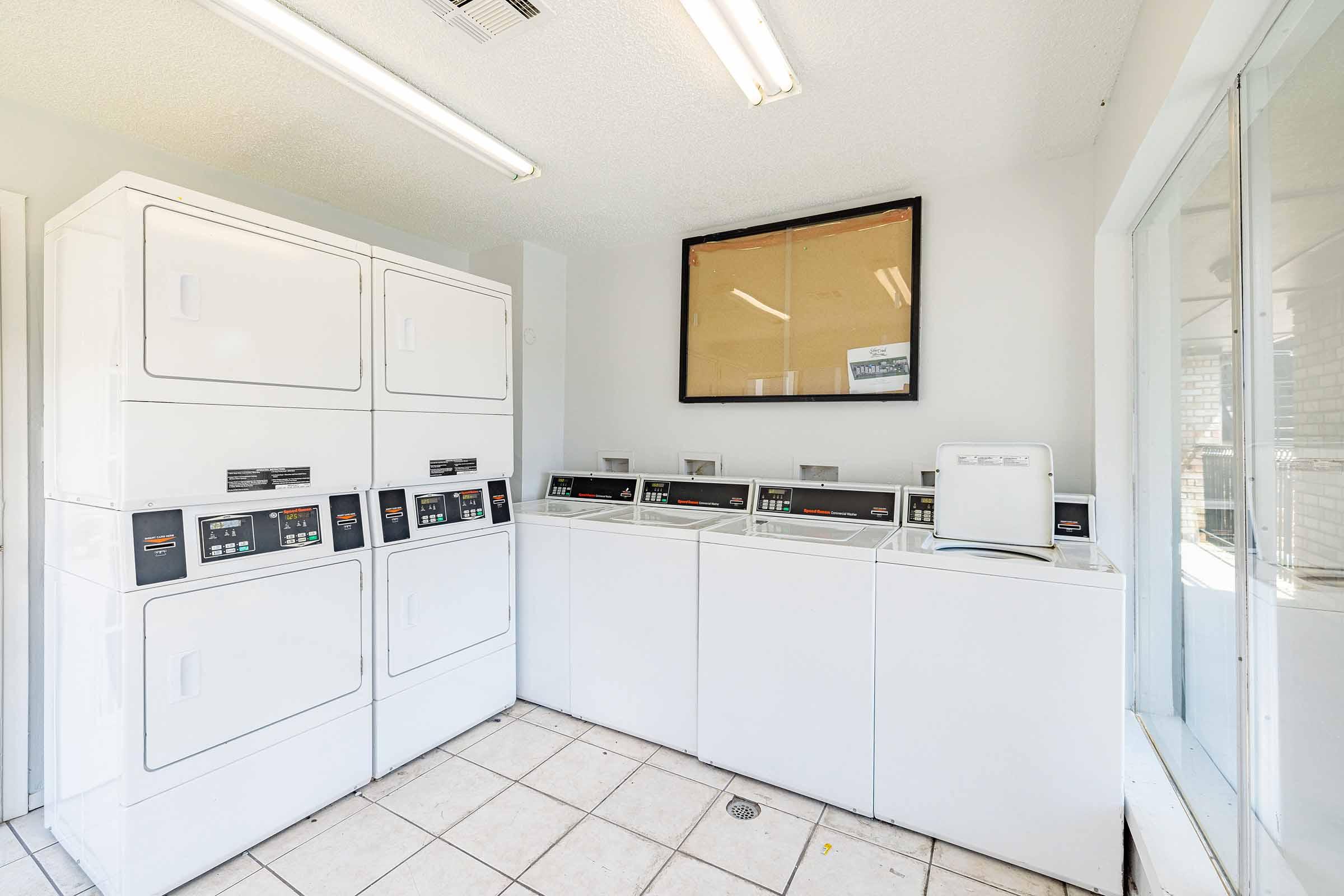 A laundry room featuring multiple white washing machines and dryers lined up against a wall. The floor is tiled, and the ceiling has bright fluorescent lights. There is a large window on one side and a framed blank notice board hanging on the wall.