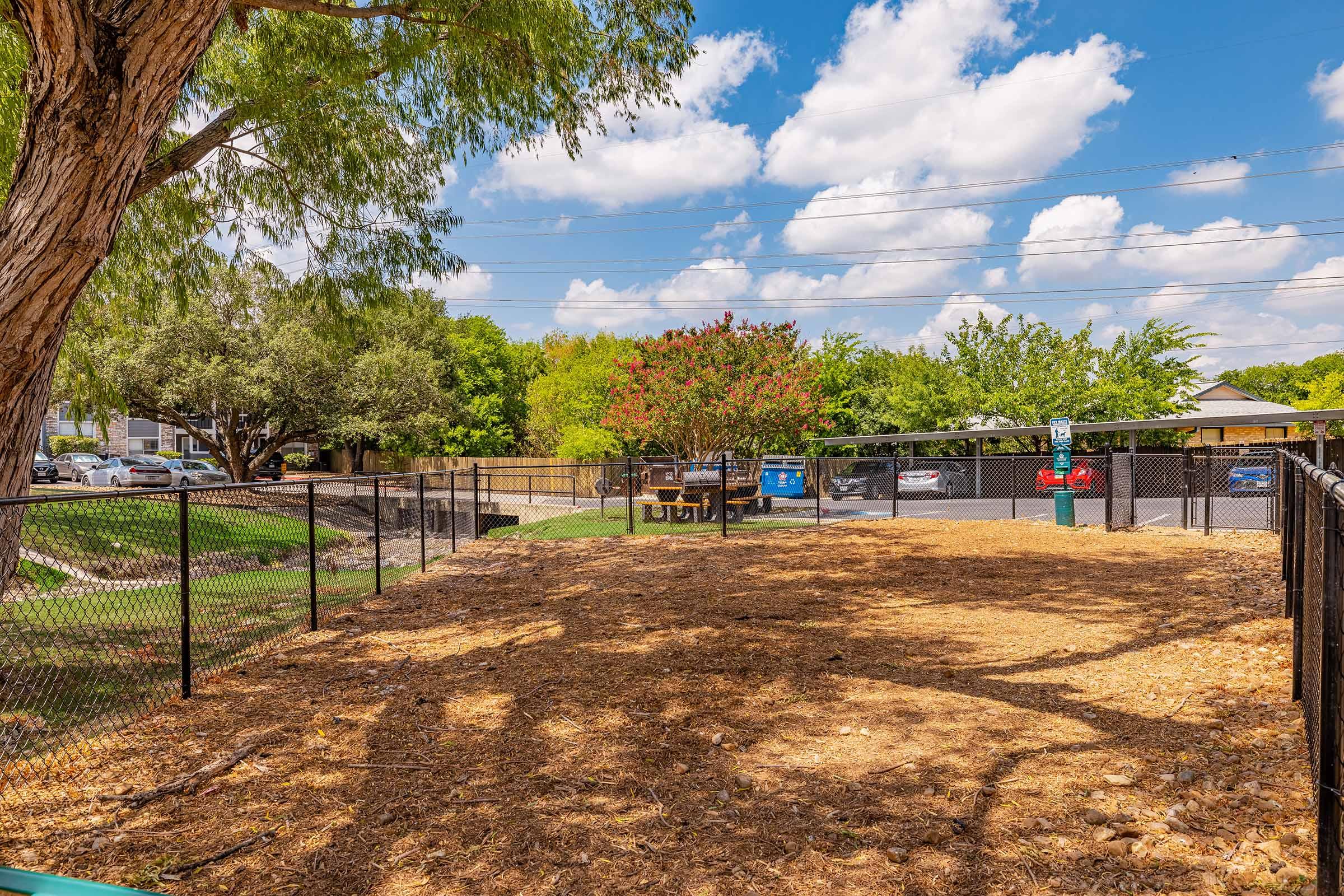 A fenced dog park with a woodchip ground, shaded by a large tree. In the background, there are green trees and parked cars, along with a blue dumpster. The sky is bright with scattered clouds. The park appears inviting and well-maintained.