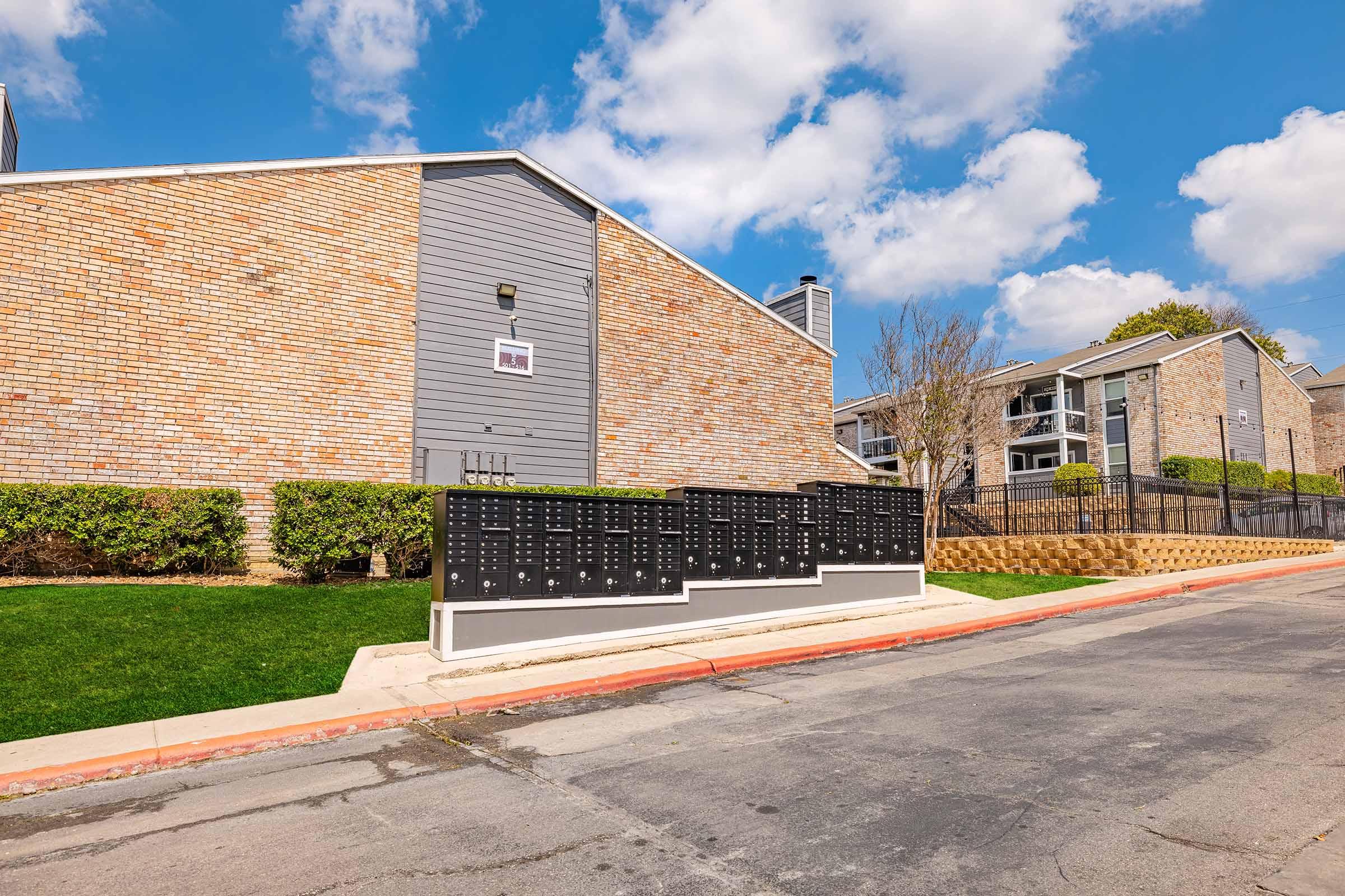 A row of black mailboxes in front of a stone and gray building, with manicured greenery along the sidewalk. The sky is partly cloudy, and the scene is well-lit, indicating a clear day. The mailboxes are arranged neatly on a concrete base near the street.