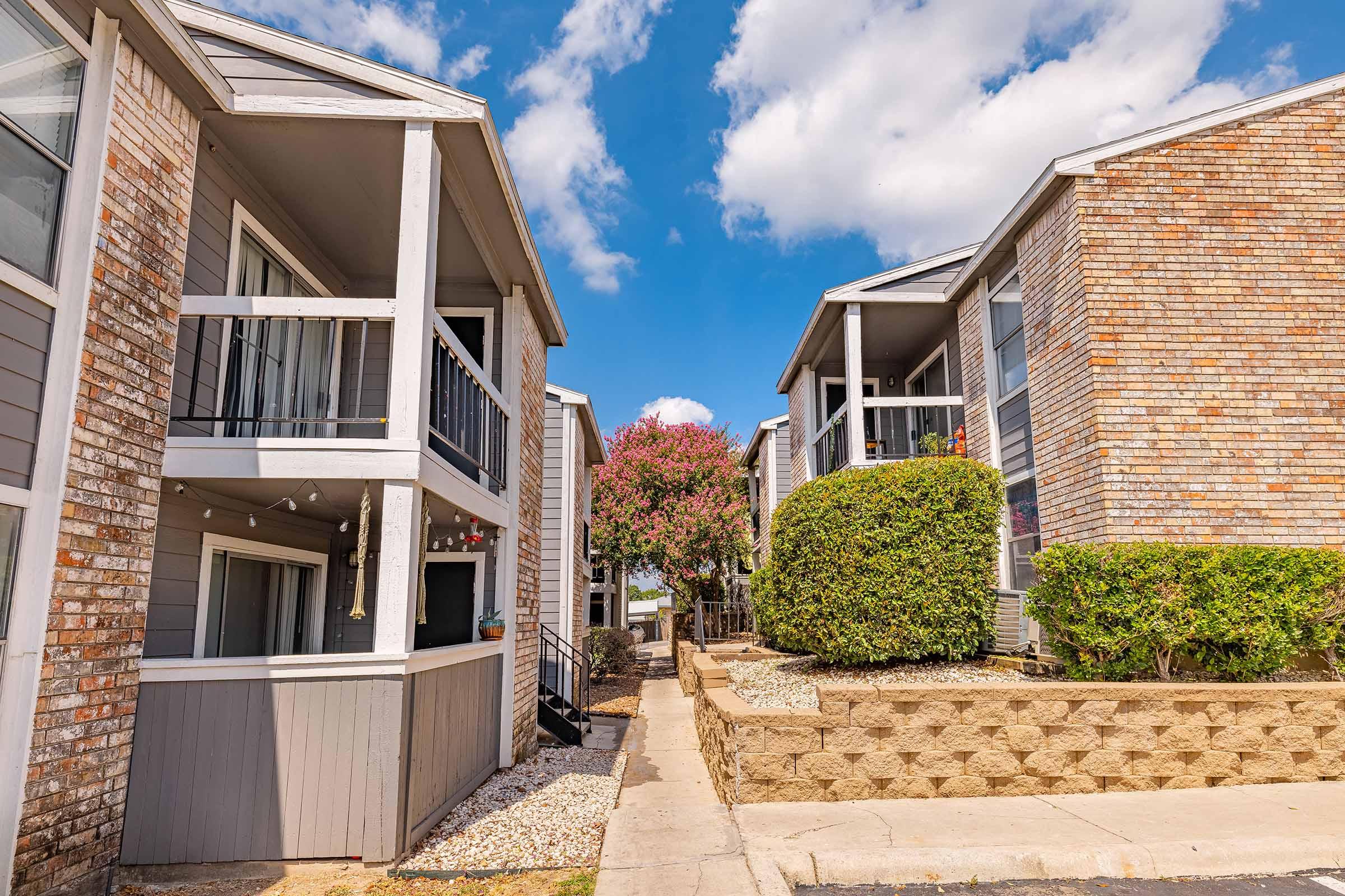 View of two apartment buildings with balconies, separated by a walkway. Lush green landscaping and small trees are visible, along with blue skies and fluffy white clouds. The buildings feature brick and gray exteriors, creating a pleasant residential atmosphere.