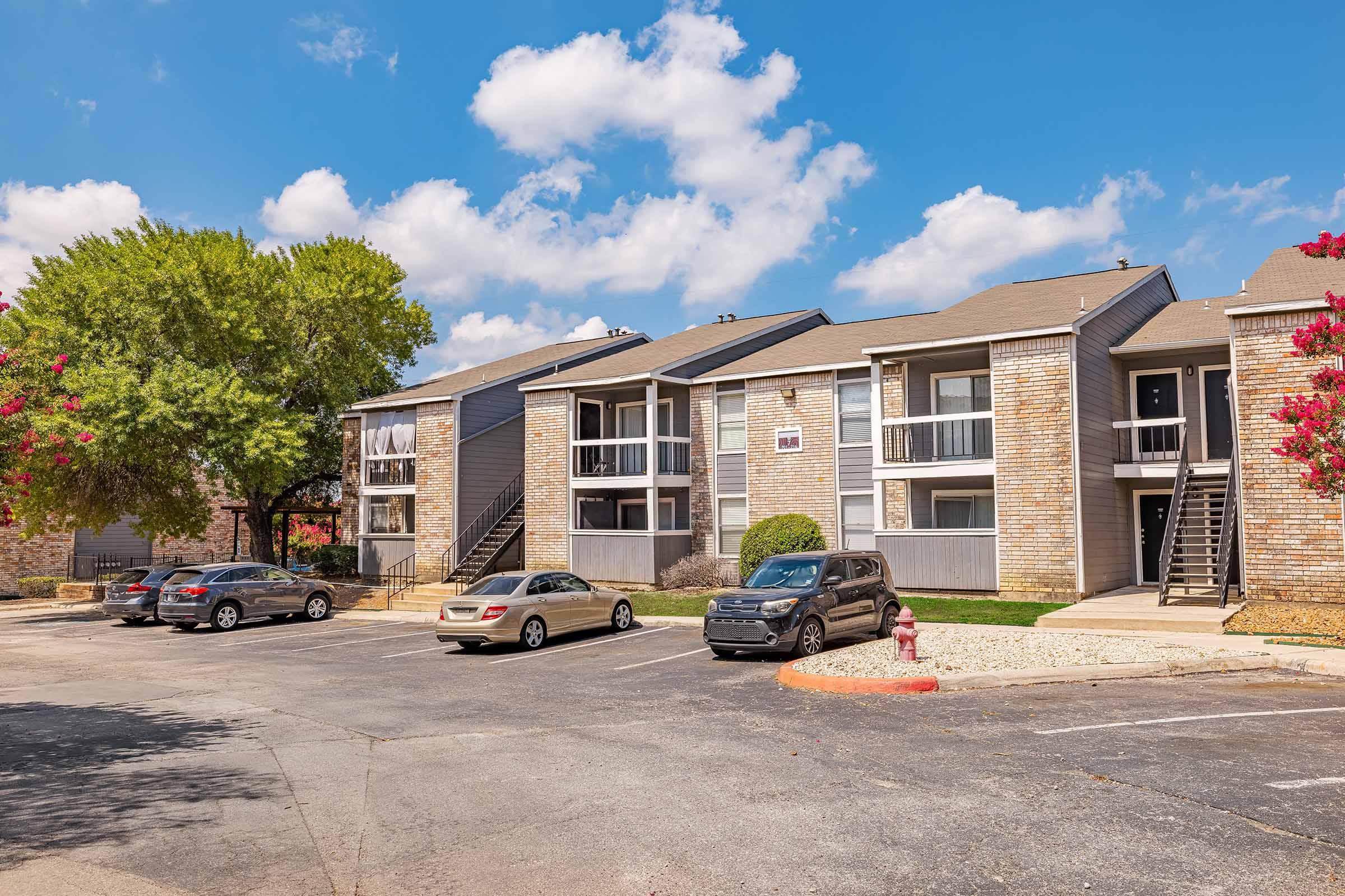 A multi-story apartment complex with brick and siding exteriors, surrounded by green trees and shrubs. The parking lot in front has several cars parked, and the sky is partly cloudy with blue and white clouds. A fire hydrant and a landscaped area are visible in the foreground.