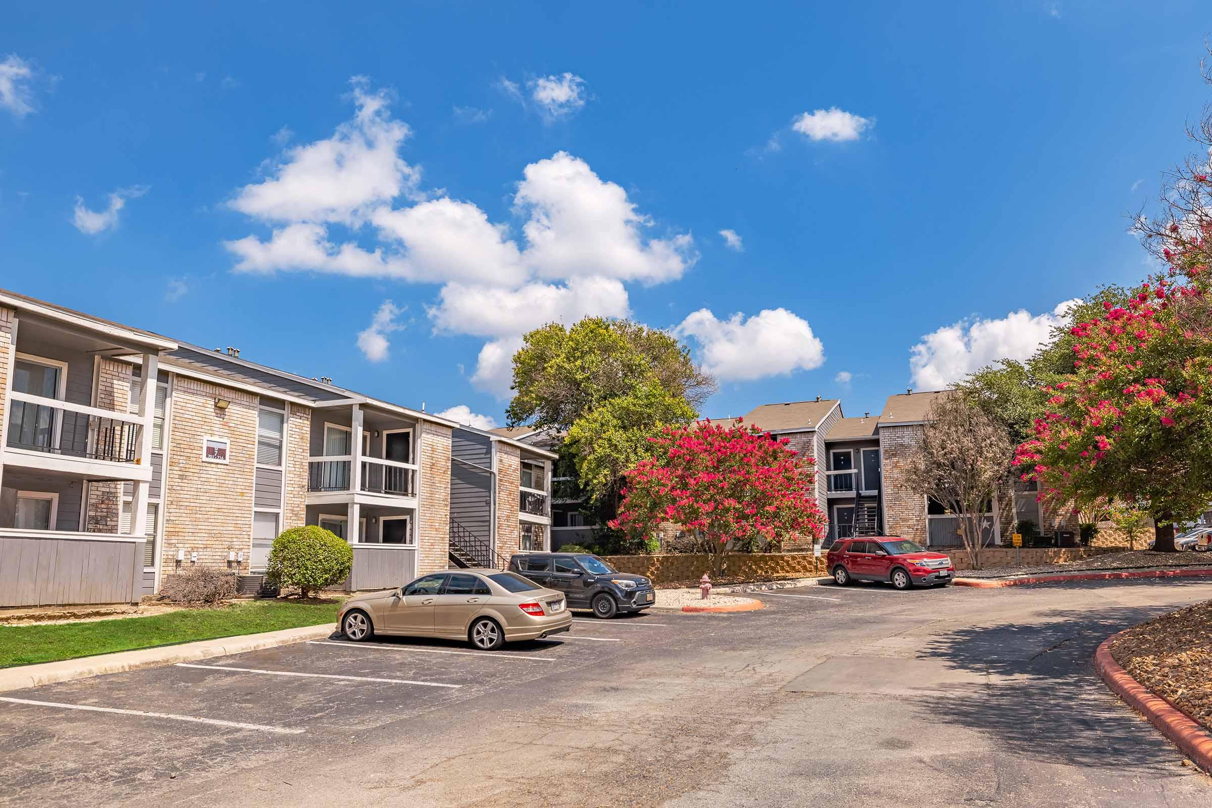A view of an apartment complex featuring several two-story buildings, a parking area with parked cars, and vibrant pink flowering trees. The sky is bright with scattered clouds, creating a pleasant atmosphere.
