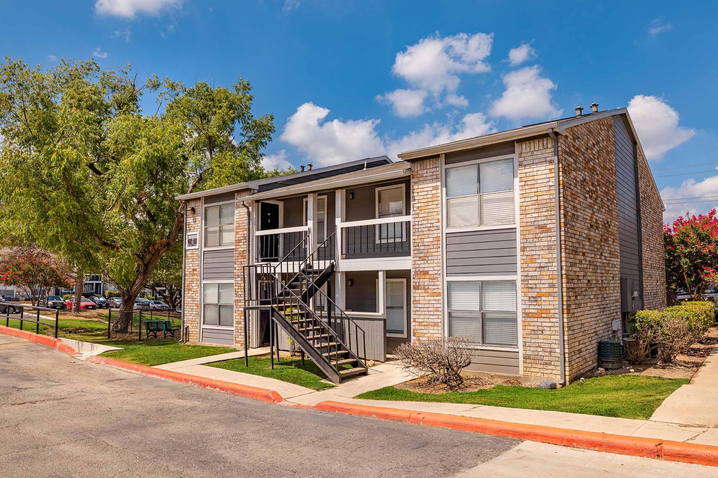 Two-story apartment building with a brick exterior featuring a staircase leading to the second floor. Lush green lawn and trees surround the building, with a clear blue sky and scattered clouds in the background. A paved path runs along the front of the structure.