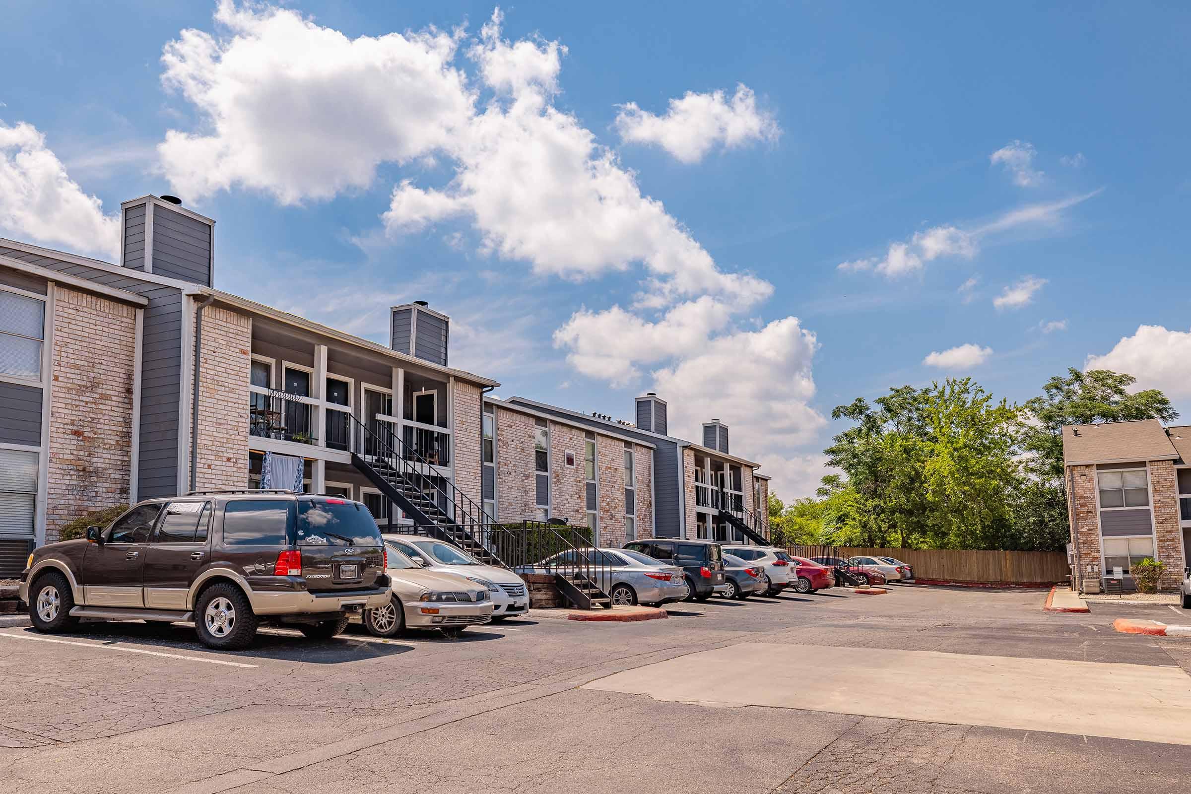 A residential apartment complex featuring two-story buildings with balconies, surrounded by a parking lot. Various parked cars are visible, and the scene includes blue skies with fluffy clouds and trees in the background.