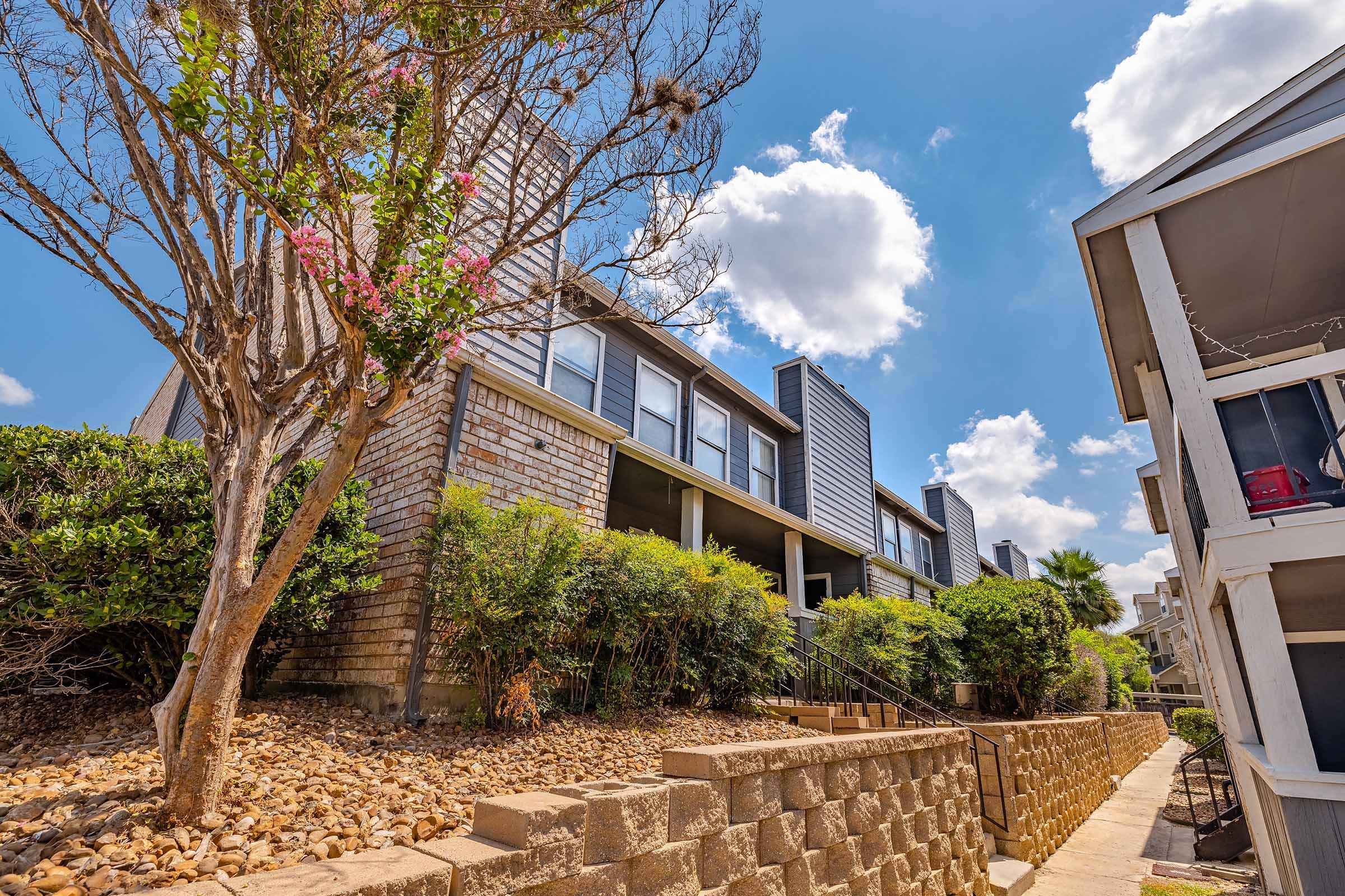 A view of a residential area featuring a multi-level building with stone and wooden exterior. The scene includes a tree with pink flowers in the foreground, landscaped shrubbery, and a staircase leading up to the building. The sky is bright blue with fluffy white clouds.