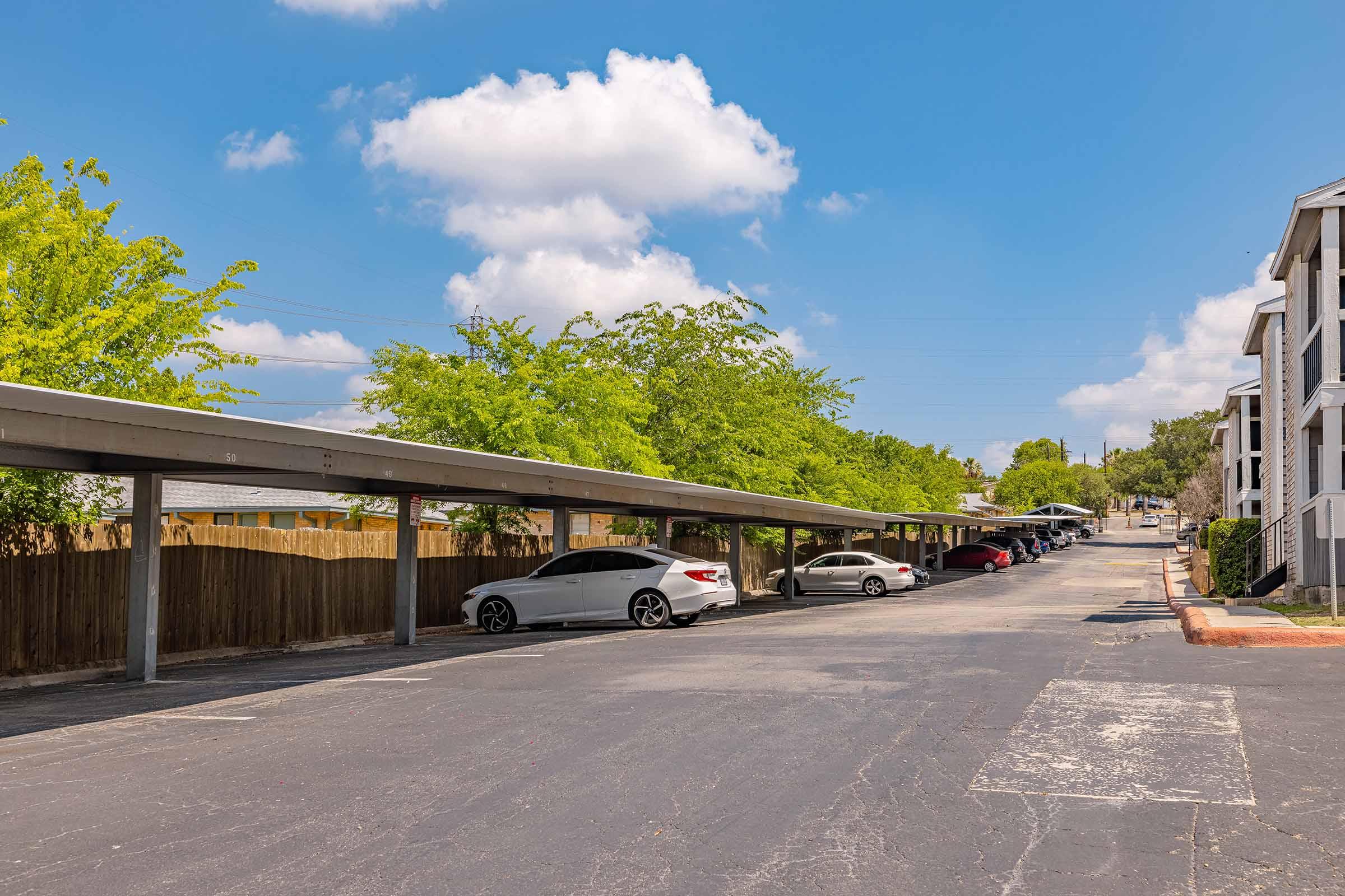 A sunny day in a parking lot with covered spaces. Several cars are parked under the shade, with some greenery in the background. The sky is blue with a few fluffy clouds, and there's a row of apartment buildings in the distance. The paved lot shows signs of wear.