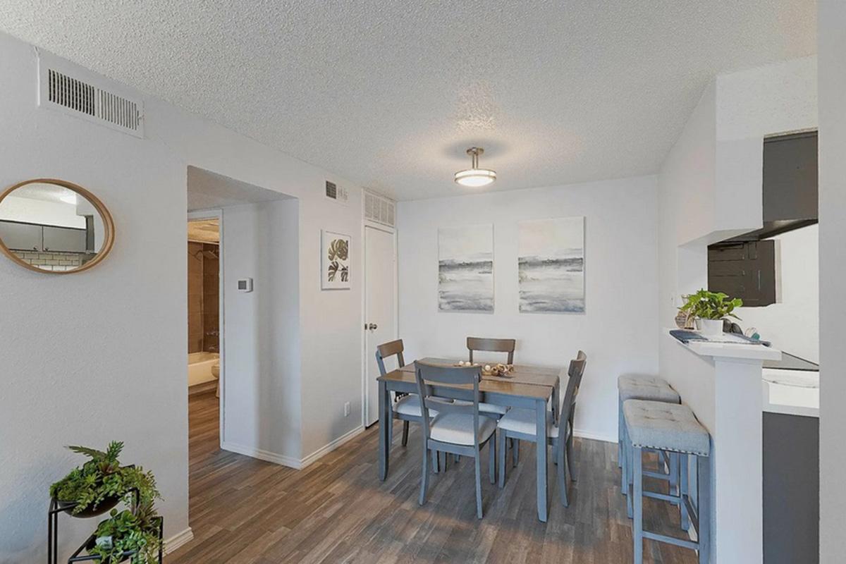 A bright and modern dining area featuring a wooden table with four chairs, a circular mirror on the wall, and two abstract paintings. The space has a light color palette and is well-lit by a ceiling fixture. In the background, there is a small kitchenette with bar seating and a view into another room.