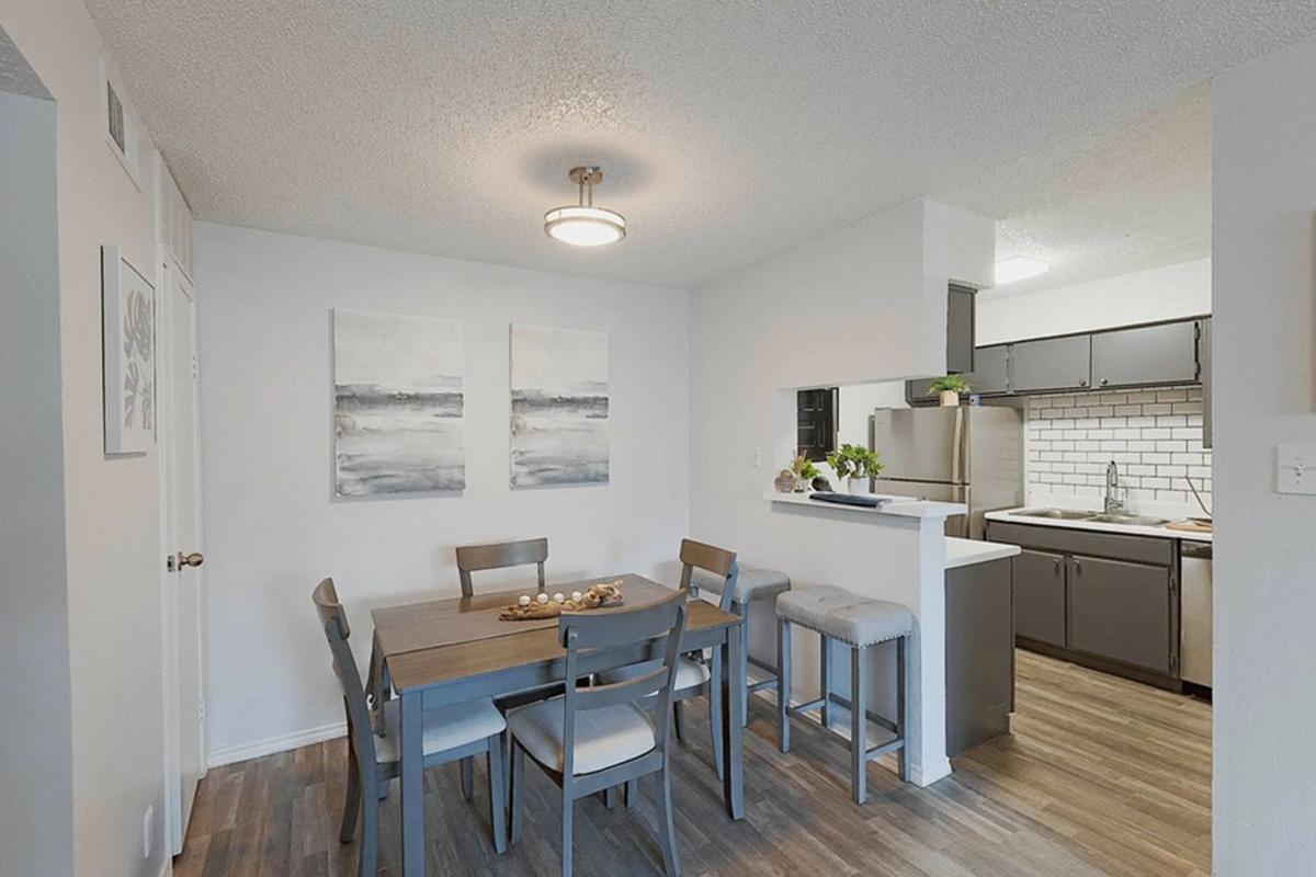 Modern dining area with a wooden table surrounded by six chairs. Two abstract paintings on the wall add a touch of decor. A sleek kitchen with dark cabinets and white subway tile can be seen in the background. Natural light illuminates the space, creating a cozy atmosphere.