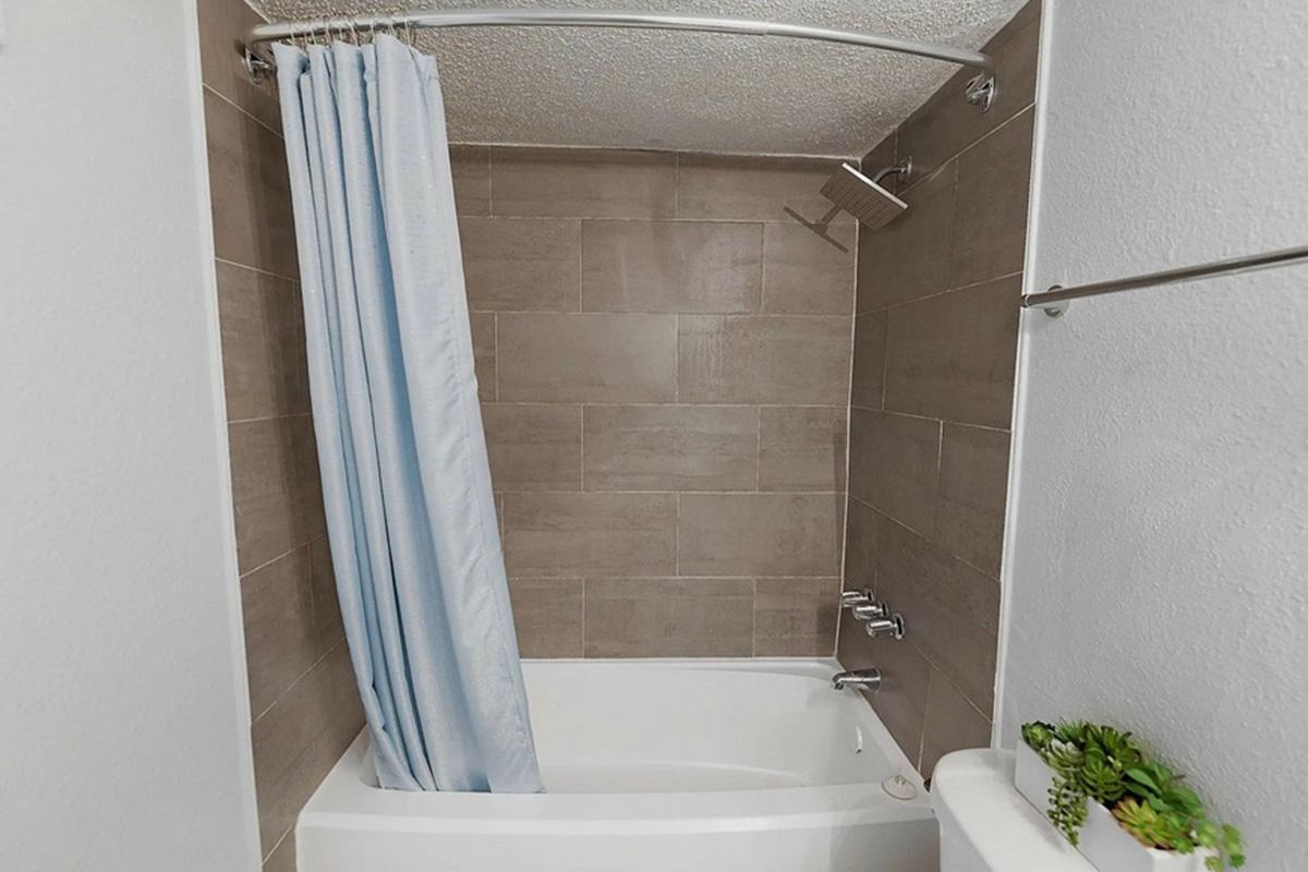 A clean and modern bathroom featuring a white bathtub with a light blue shower curtain. The walls are covered in brown tiles, and a small plant is placed on the side. The ceiling is textured, and there are several shower fixtures visible.