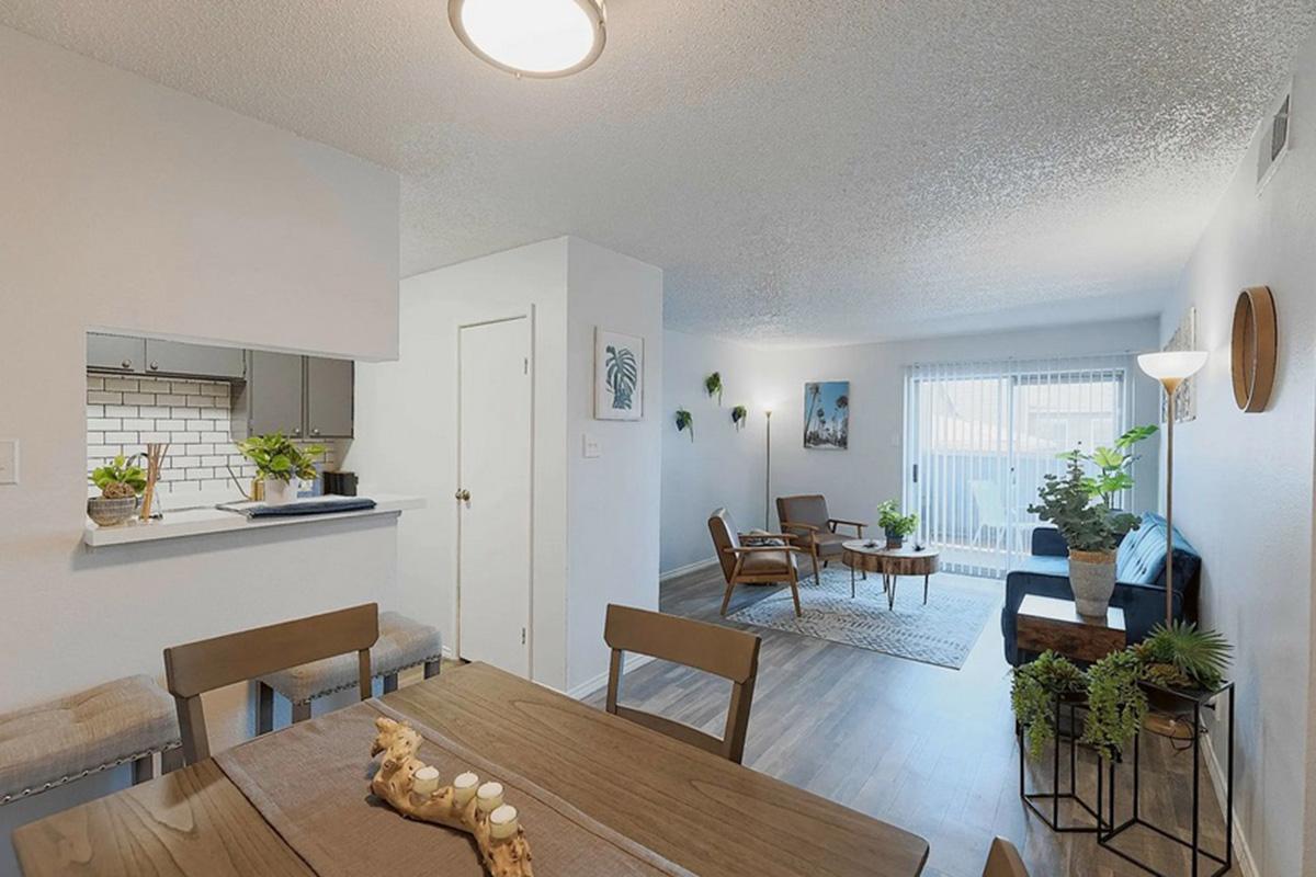 A cozy, modern living space featuring a dining area with a wooden table and chairs, a small kitchen in the background, and a bright living room with a blue sofa, plants, and a rug. Large windows allow natural light, enhancing the inviting atmosphere.