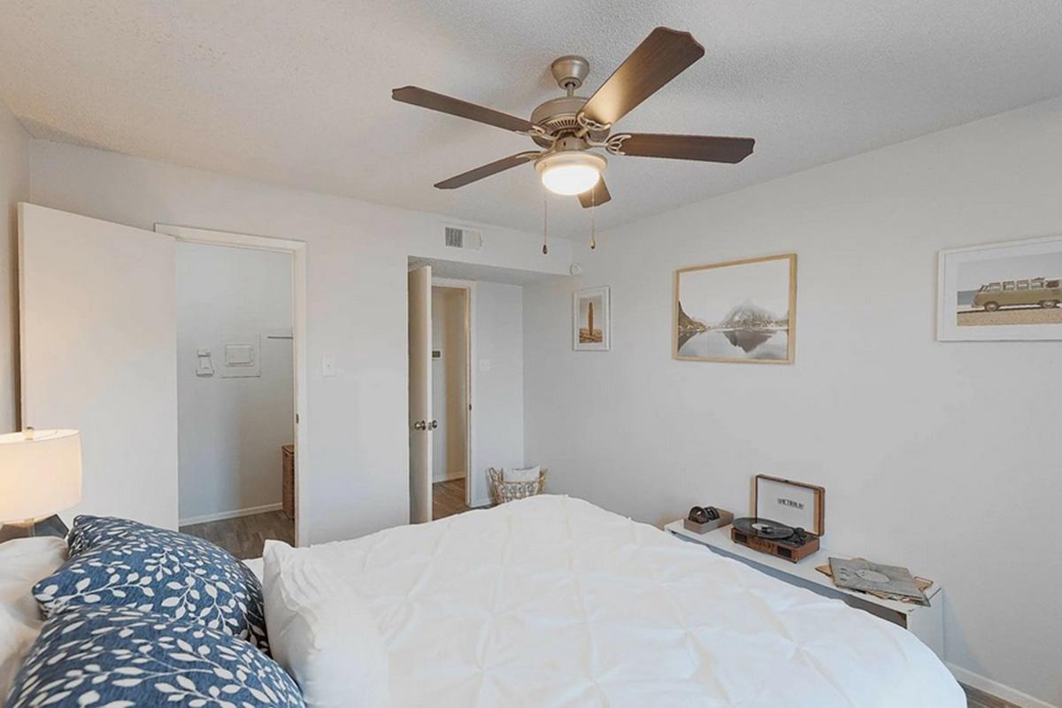 A cozy bedroom featuring a white bed with a patterned blue and white duvet, a ceiling fan, and minimalistic decor. There are framed pictures on the wall, a small side table with a vintage record player, and an open door leading to a bathroom. The overall ambiance is inviting and well-organized.