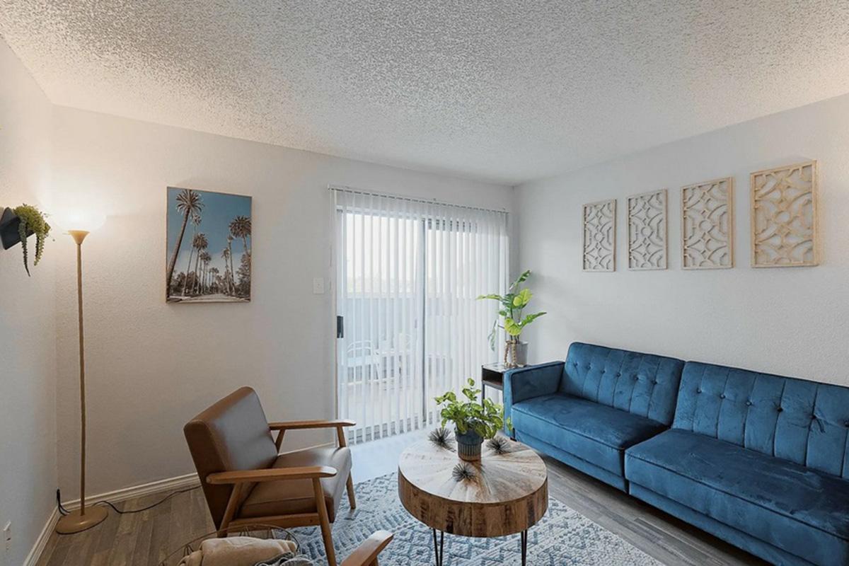 A cozy living room featuring a blue sofa, a wooden armchair, and a round coffee table with plants. There are light-colored walls with decorative wall art and large windows covered by white blinds, letting in natural light. A floor lamp adds additional warmth to the space.