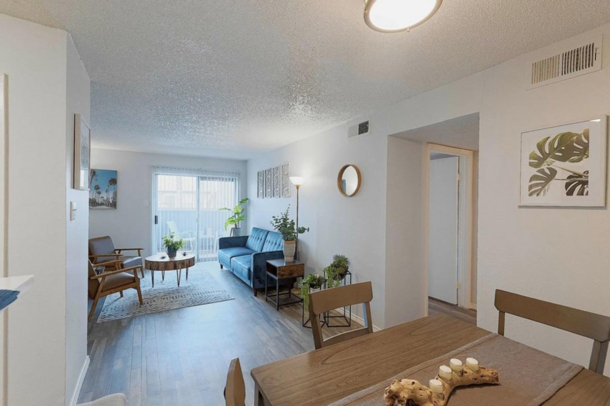 A modern living space featuring a cozy seating area with a blue sofa, wooden chairs, and indoor plants. There is a dining table with decorative items, and the walls are adorned with framed artwork. Natural light fills the room, creating a welcoming atmosphere.
