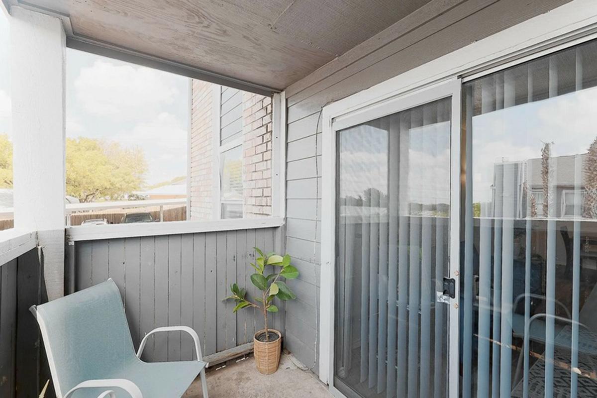 A small balcony featuring a light blue chair, a potted plant, and sliding glass doors. The walls are painted gray, with a light-colored ceiling and a view of trees outside. The space is cozy and inviting, perfect for relaxation.