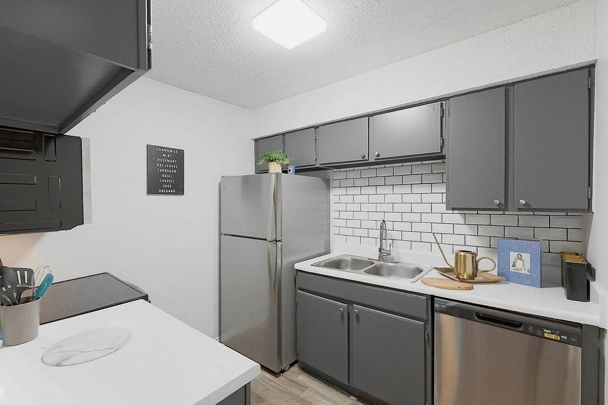A modern kitchen featuring gray cabinets, a stainless steel refrigerator, and a double sink. The countertop is white with a cutting board and a cup. A black and white framed print hangs on the wall, and there is a stove and dishwasher visible. The overall design is sleek and contemporary.