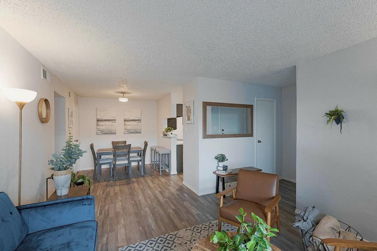 Cozy living space featuring a blue sofa, a wooden coffee table with plants, a dining area with a table and chairs, and a kitchenette in the background. The decor includes modern artwork on the walls, a floor lamp, and a decorative wall hanging, creating a warm and inviting atmosphere.