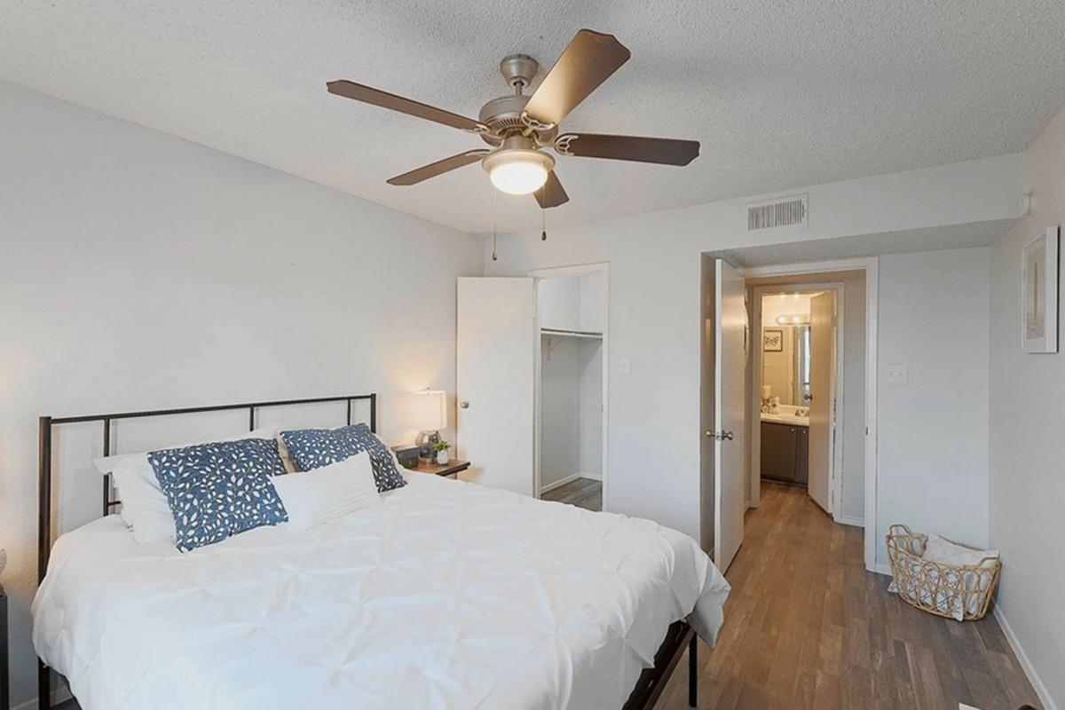 A bright, inviting bedroom featuring a queen-sized bed with white bedding and blue accent pillows. A ceiling fan hangs above, and there is a closet on the left. The room leads to a bathroom visible in the background, with neutral walls and wood-style flooring throughout. A decorative basket is in the corner.