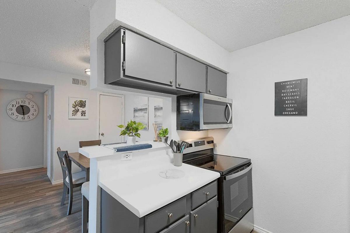 A modern kitchen featuring gray cabinets, a white countertop, and stainless steel appliances. A small dining table with chairs is visible in the background. The wall has a decorative sign with a list of food items, and a clock is displayed nearby. Bright lighting enhances the clean and inviting atmosphere.