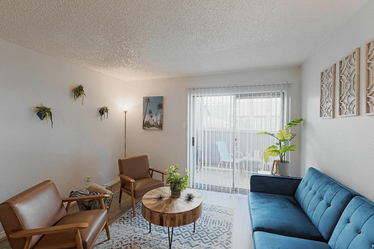 A cozy living room featuring a blue sofa, two brown armchairs, and a round coffee table with a plant. The walls have decorative accents and small indoor plants. Large sliding doors open to a balcony with outdoor seating, allowing natural light to fill the space.