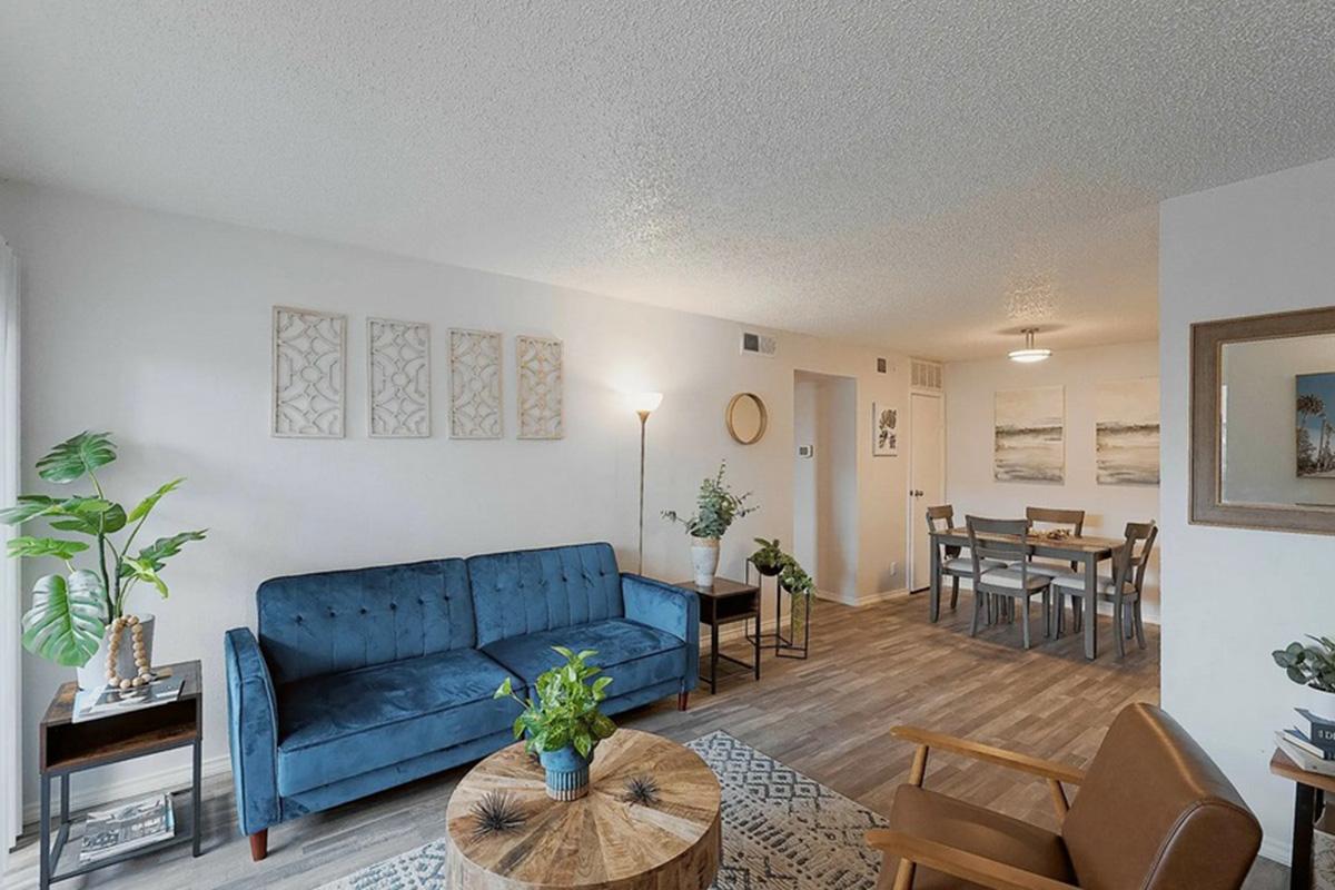 A cozy living room featuring a blue velvet sofa, a wooden coffee table, and a stylish lamp. The space has light-colored walls adorned with framed art. A dining area with a wooden table and chairs is visible, along with plants adding a touch of greenery, creating a warm and inviting atmosphere.