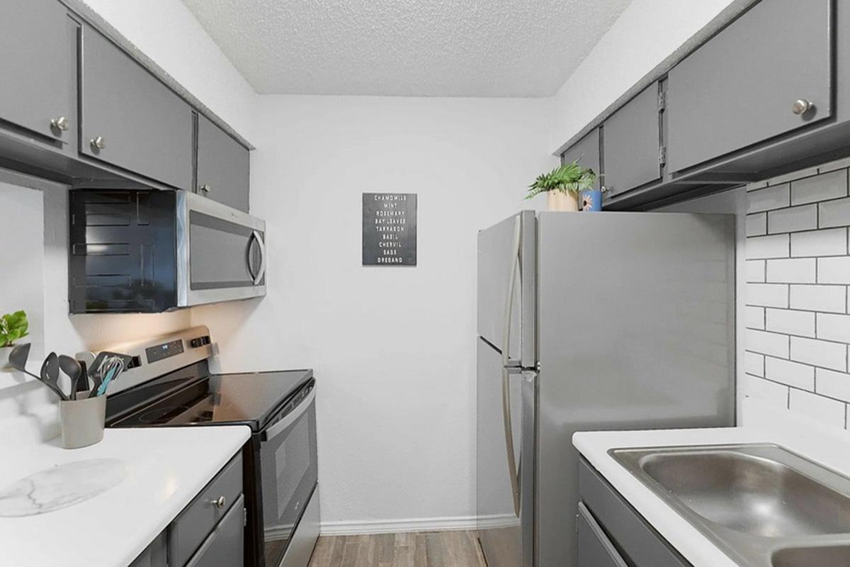A modern kitchen featuring gray cabinets, stainless steel appliances, and a white tile backsplash. The space includes an oven, microwave, refrigerator, and a sink, with a small wall decoration above the counter. The floor is light wood, and there are green plants adding a touch of color.