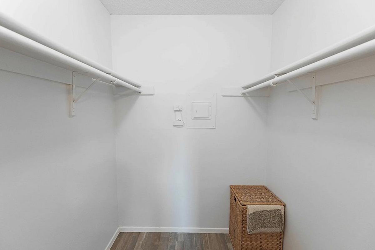 Empty closet with two white hanging rods and a wicker storage basket on the floor. The walls are plain white, and the flooring is a light-colored wood. A small panel is visible on the wall, and the overall space is bright and uncluttered.