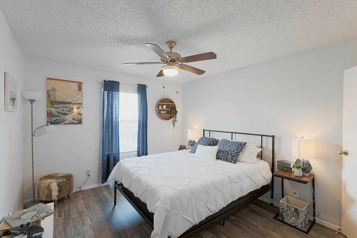 A cozy bedroom featuring a queen-sized bed with a white duvet, two decorative pillows, and bedside lamps. There's a ceiling fan and a wall-mounted art piece. A window with blue curtains lets in natural light. A small side table and a decorative storage basket add to the room's aesthetic.