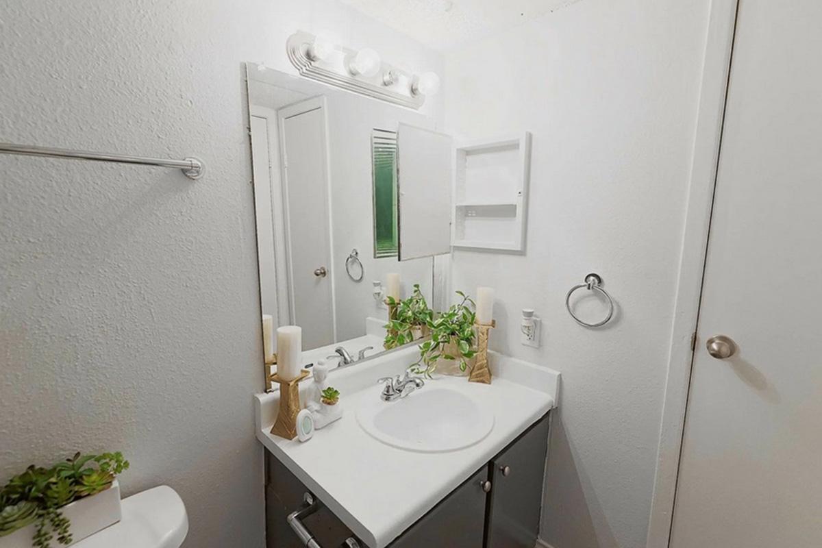 A small bathroom featuring a white sink and countertop, a rectangular mirror above the sink, and a light fixture with multiple bulbs. The walls are painted light gray, and there are minimal decorative elements, including a small plant and candles on the countertop. A towel rack is mounted on the wall.