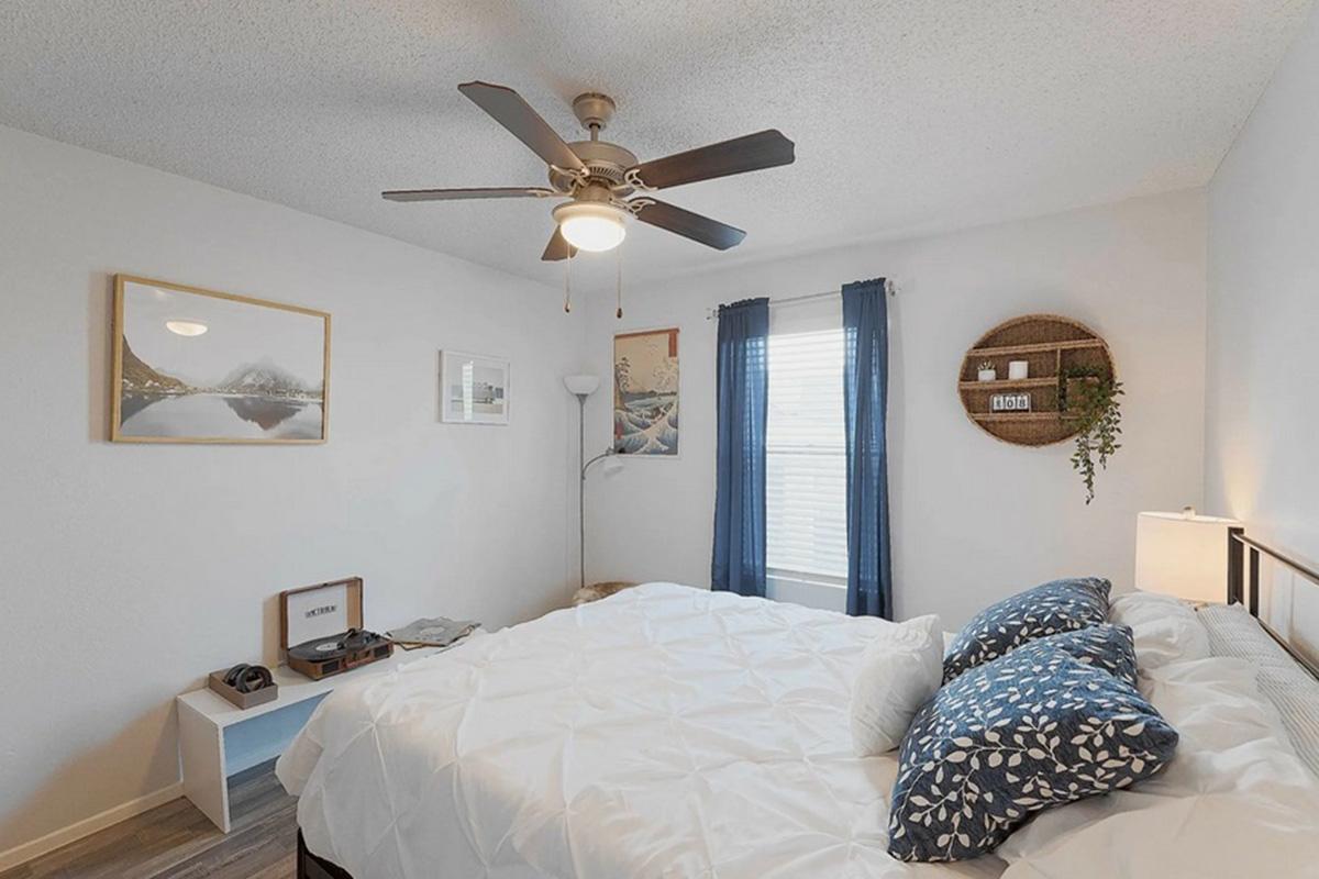 Cozy bedroom featuring a queen-sized bed with decorative blue and white pillows, a ceiling fan, and two windows with blue curtains. A small nightstand holds a lamp and decor, while a wall-mounted shelf displays items like plants and candles. Natural light brightens the space, creating a warm atmosphere.