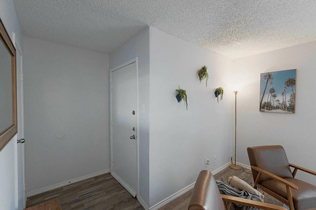 A cozy corner of a room featuring a simple entrance door, a floor lamp, and two wooden chairs. On the wall, small planters with greenery hang, adding a touch of nature. A framed photo of palm trees is mounted on the wall, enhancing the warm atmosphere of the space.