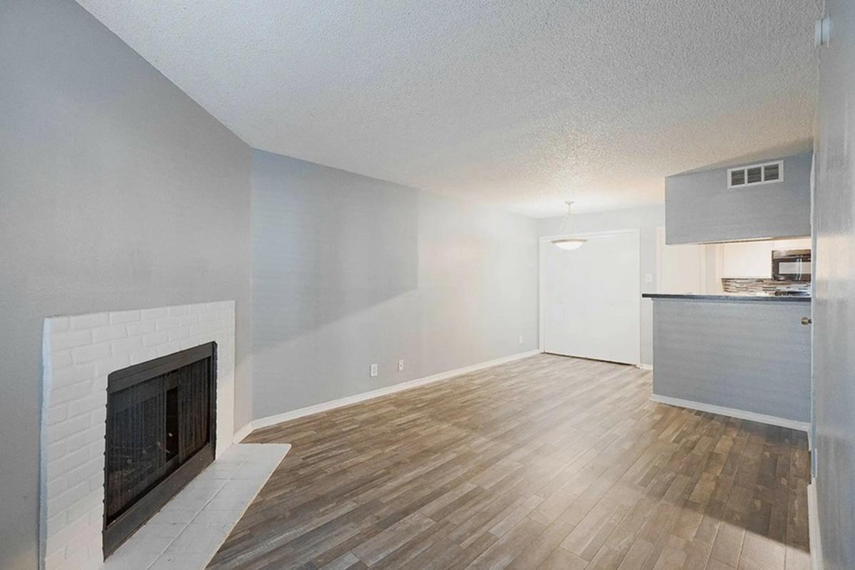 Spacious living room with light gray walls and wood-look flooring, featuring a white brick fireplace. A doorway leads to a dimly lit area, likely a kitchen. Natural light from a ceiling fixture illuminates the space. The overall atmosphere is modern and inviting.