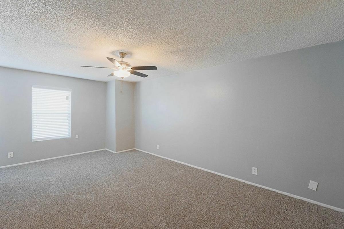 A spacious and empty room featuring light gray walls, a ceiling fan with five blades, and a window with blinds. The carpeted floor is also light gray, and there are no furnishings or decorations present, creating a clean and simple atmosphere.