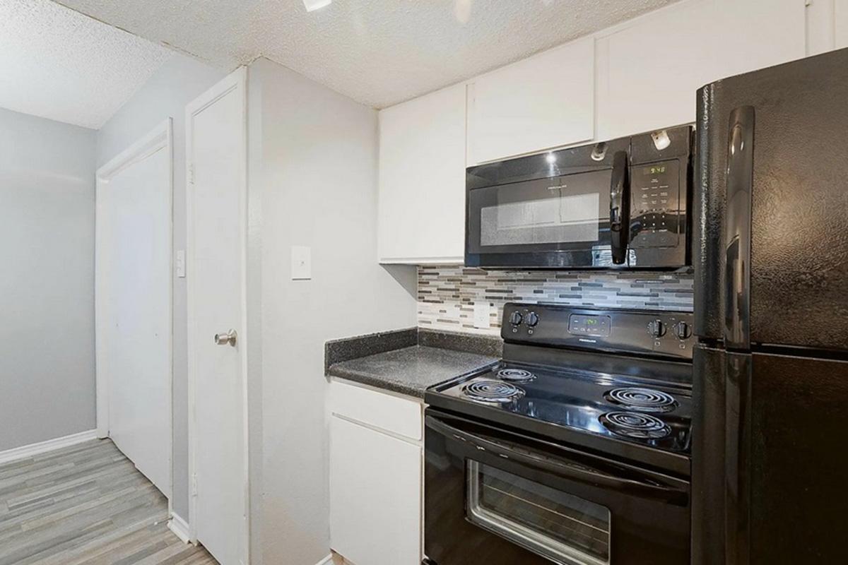 A modern kitchen featuring a black refrigerator, microwave, and stove. The countertops are dark, and the backsplash consists of small, multi-colored tiles. Light gray walls and a wooden floor complete the space, with a door visible in the background.