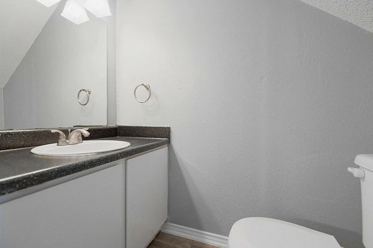 A simple bathroom interior featuring a countertop with a sink and faucet, a mirror above the sink, a towel ring, and a white toilet. The walls are painted light gray, and the flooring is wooden. The lighting comes from a ceiling fixture.