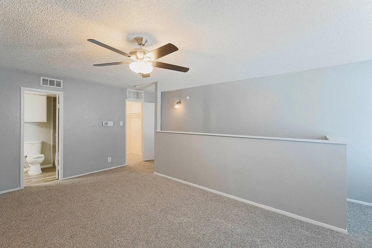 A spacious, well-lit living area featuring a ceiling fan, beige carpet, and light gray walls. To the left, a doorway leads to a bathroom with visible fixtures. The layout includes a divider wall and ample natural light from nearby windows.