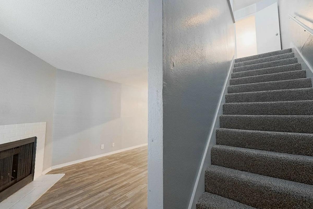 A view of a residential interior showing a staircase leading to a brightly lit area. The left side features a cozy living space with a fireplace and grey walls, while the right side displays a well-lit hallway at the top of the stairs. The floor has a mix of wood-like and carpeted surfaces.