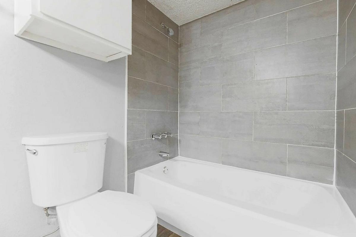 A modern bathroom featuring a white bathtub and toilet with gray tiled walls. The space is well-lit and has a clean, minimalist design. A small shelf is mounted above the toilet.