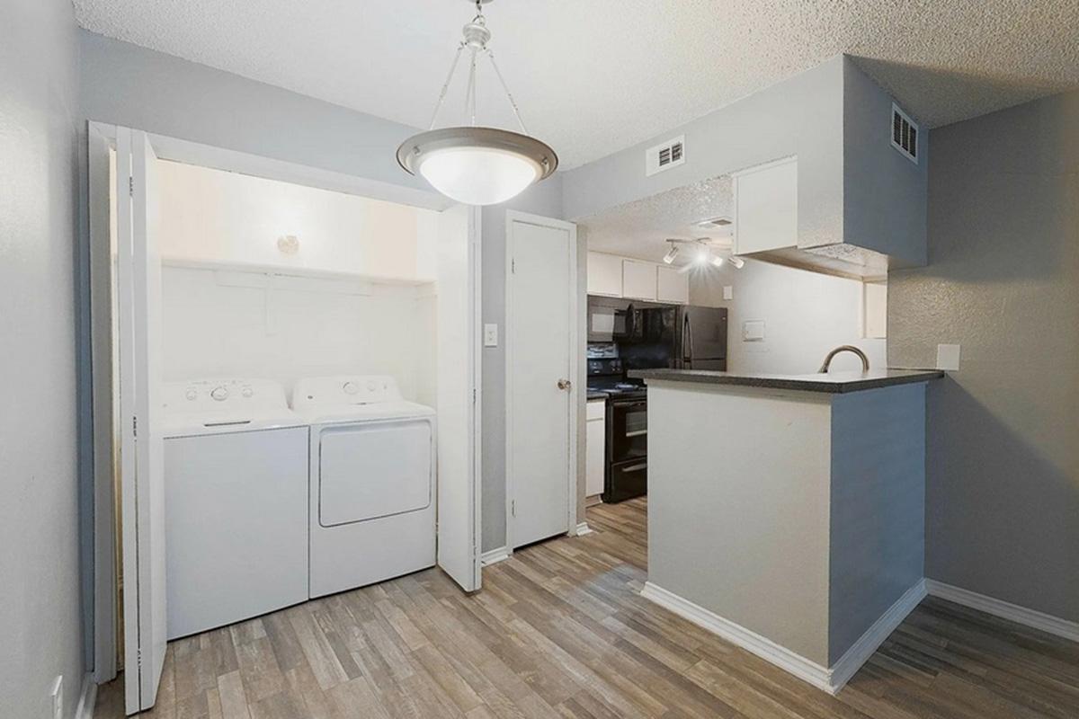 A view of a modern laundry area featuring stacked washer and dryer units, with a minimalist kitchen visible in the background. The light gray walls and wood-like flooring create a contemporary look, and a pendant light hangs from the ceiling. The space is bright and well-organized.