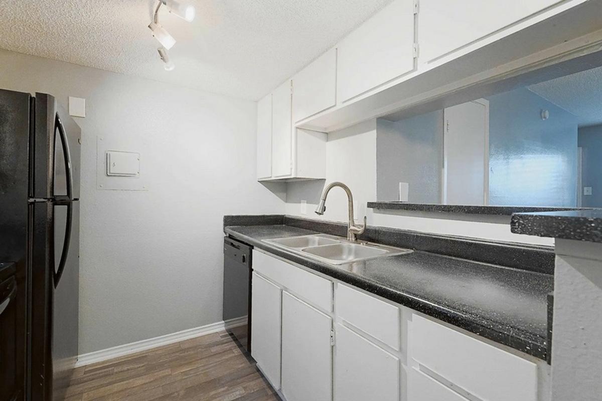 A modern kitchen featuring white cabinets, a dual sink with a chrome faucet, and black countertops. The space includes a black refrigerator and dishwasher, with hardwood-style flooring and neutral wall color, providing a clean and bright appearance. Natural light enters from an unseen window.