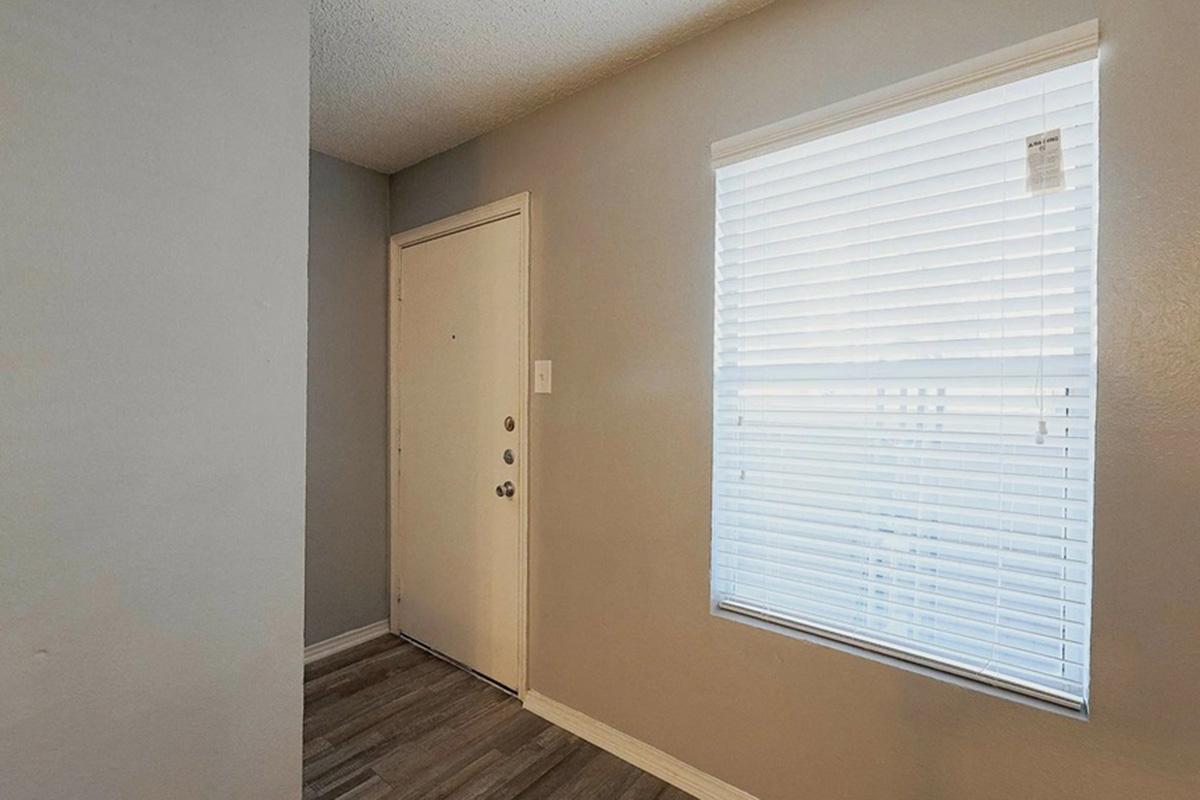 Interior view of a small entryway featuring a closed door with a doorknob and multiple locks, gray walls, and light-colored laminate flooring. A window with white blinds allows natural light into the space, and a tag is hanging from the blinds. The area appears clean and uncluttered.