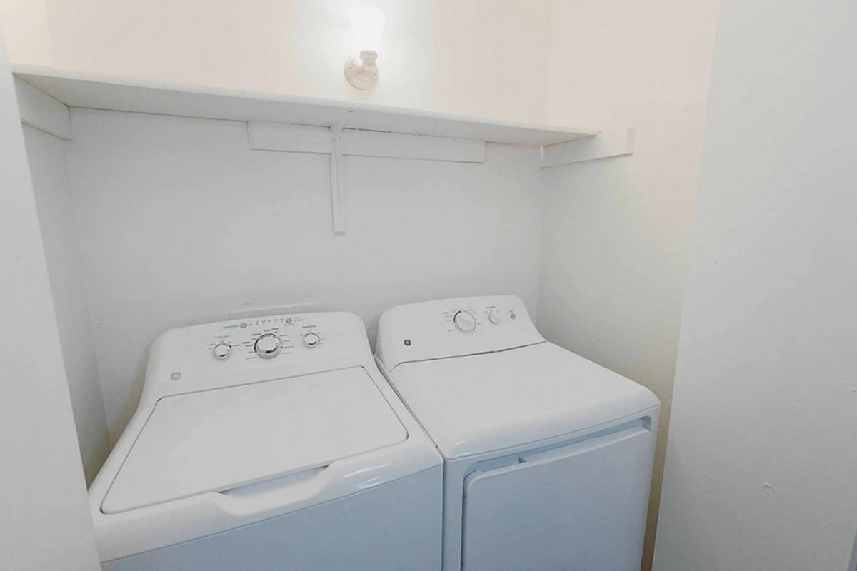 A small laundry area featuring a stacked washer and dryer against a plain white wall. There is a single overhead light fixture, and the space appears clean and uncluttered, with shelves above the appliances for additional storage.
