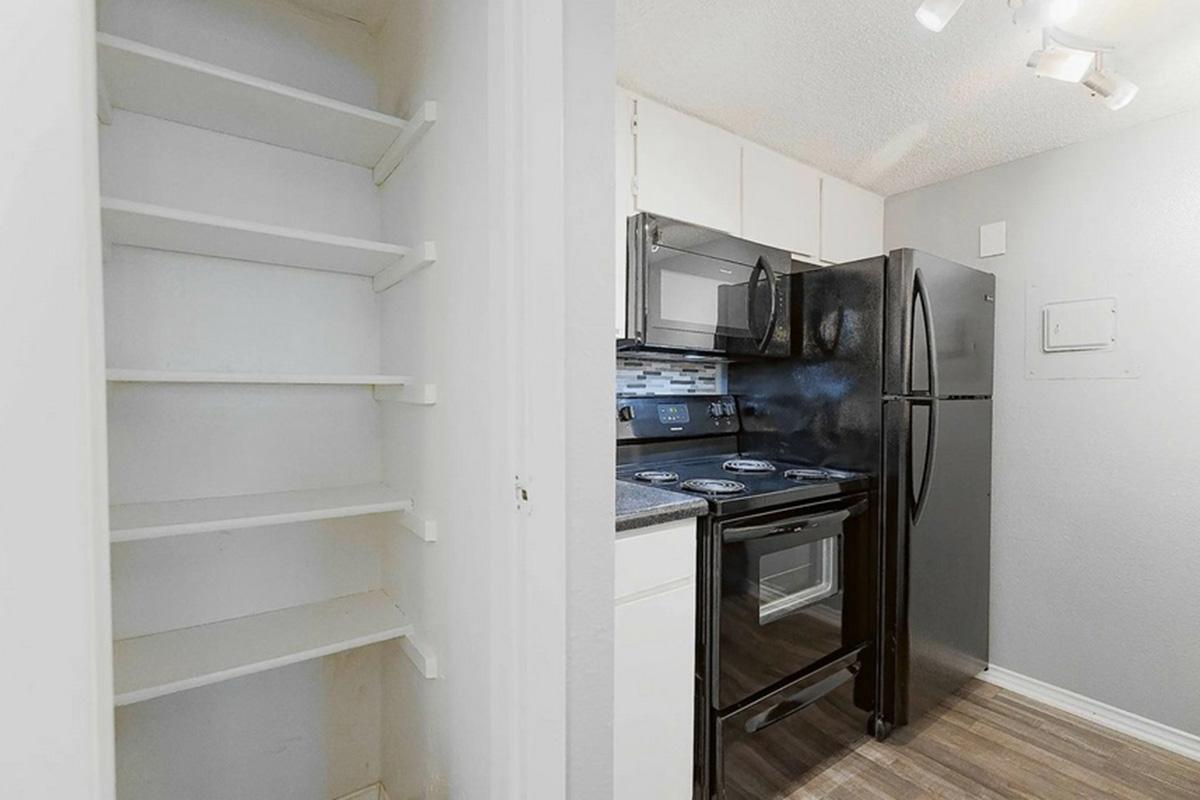 A small kitchen area featuring a black refrigerator and stove, with light-colored cabinets. To the left, there's a white shelving unit with open shelves. The floor is made of laminate wood, and the walls are painted in light tones, creating a cozy and functional space.